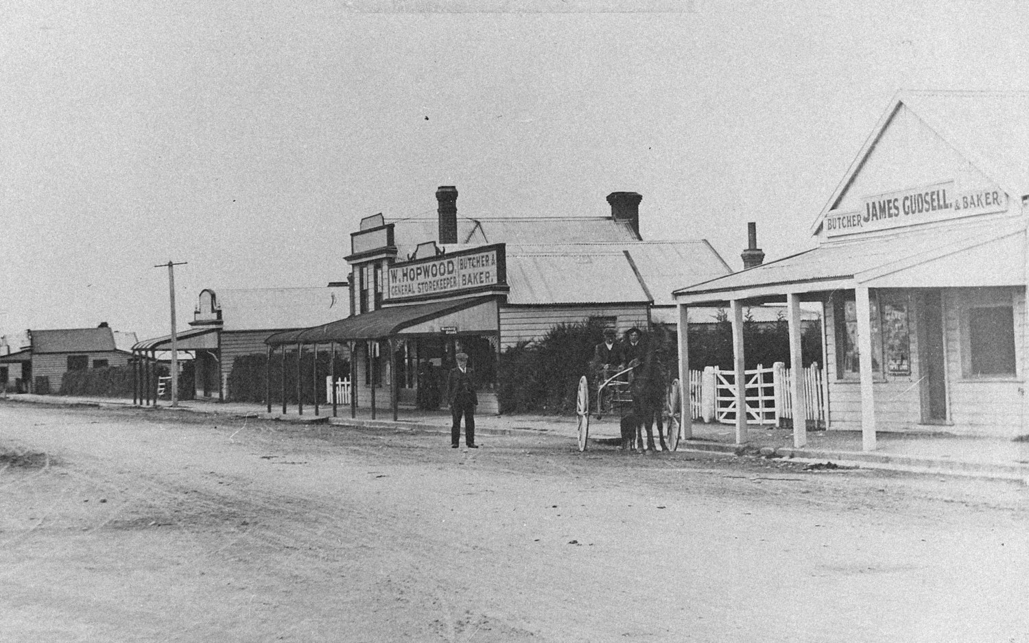 Black-and-white view along Archibald Street, Tinwald, showing a row of early shops with verandahs beside a wide unsealed road.