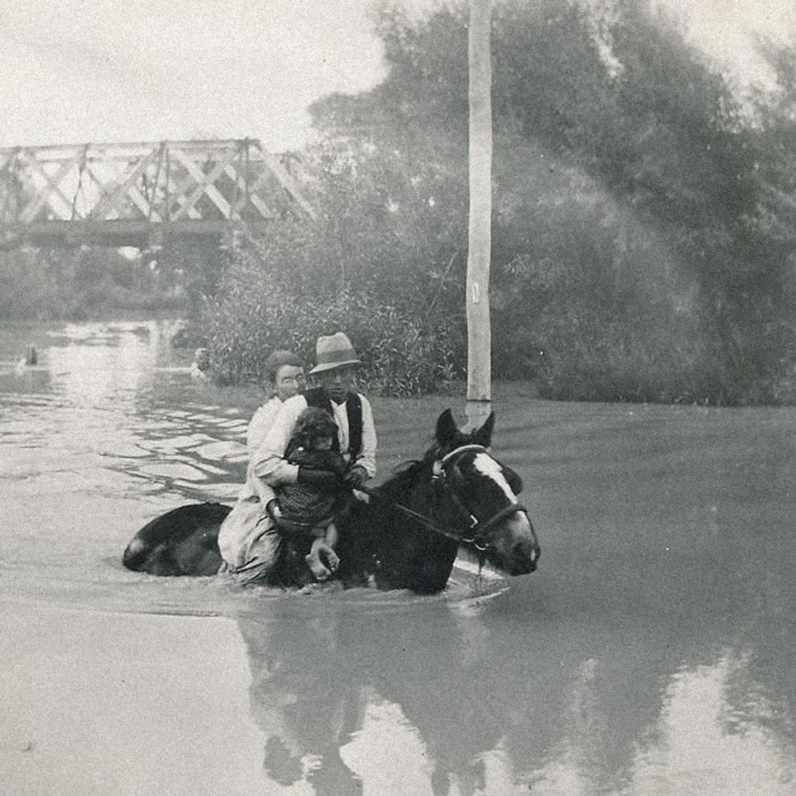 Sepia photograph of a family on horseback wading through deep floodwater, with a bridge in the background.