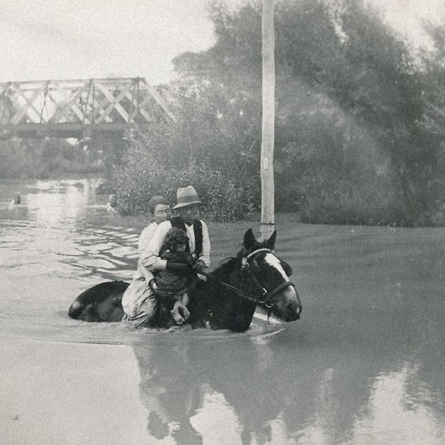 Sepia photograph of a family on horseback wading through deep floodwater, with a bridge in the background.