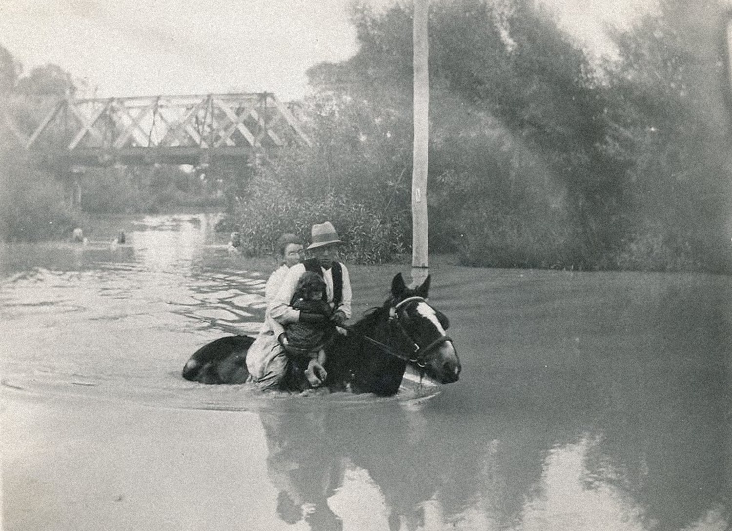 Sepia photograph of a family on horseback wading through deep floodwater, with a bridge in the background.