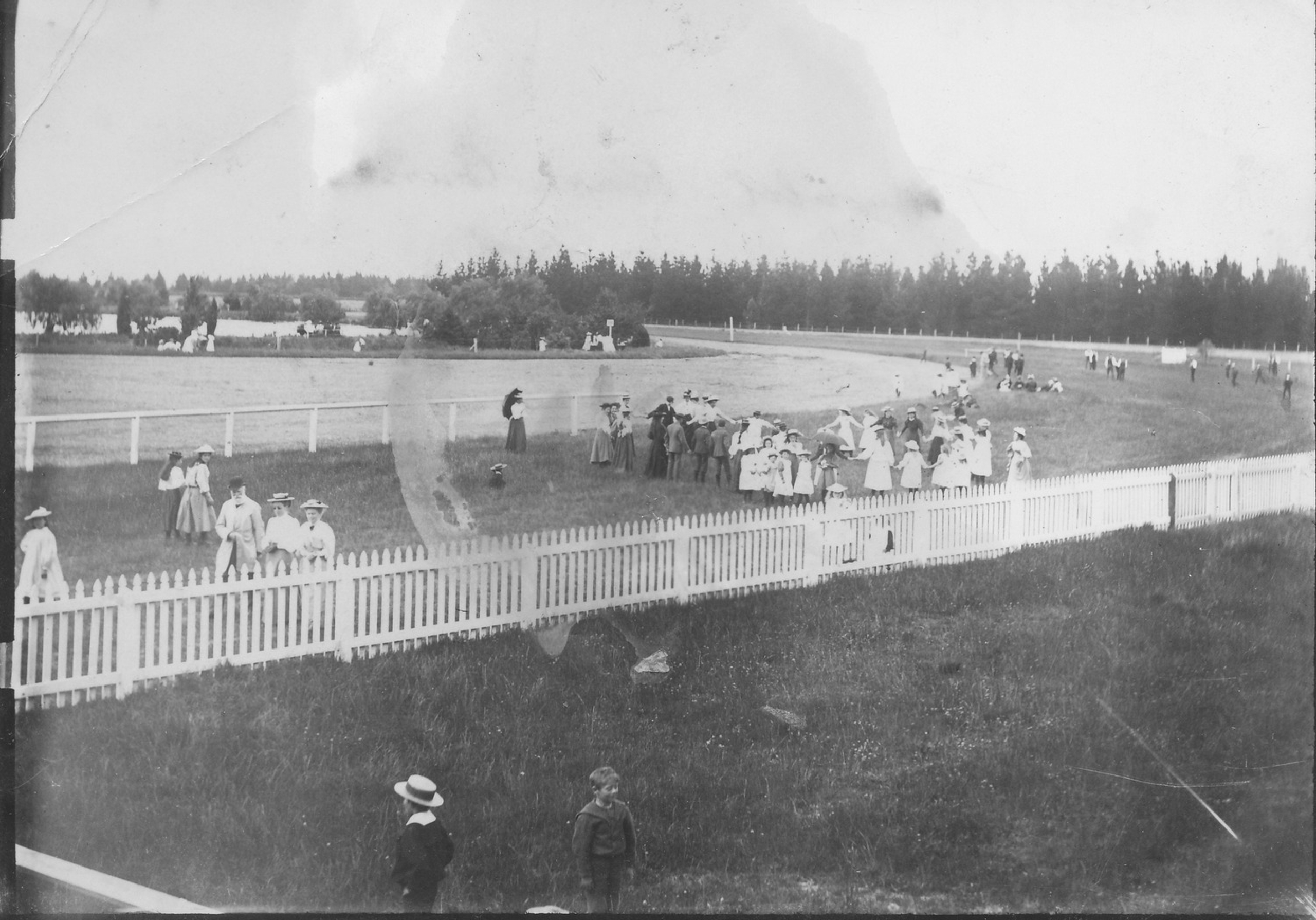 Black-and-white photograph of crowds in their best clothes gathered along the white rail of the Tinwald racecourse.