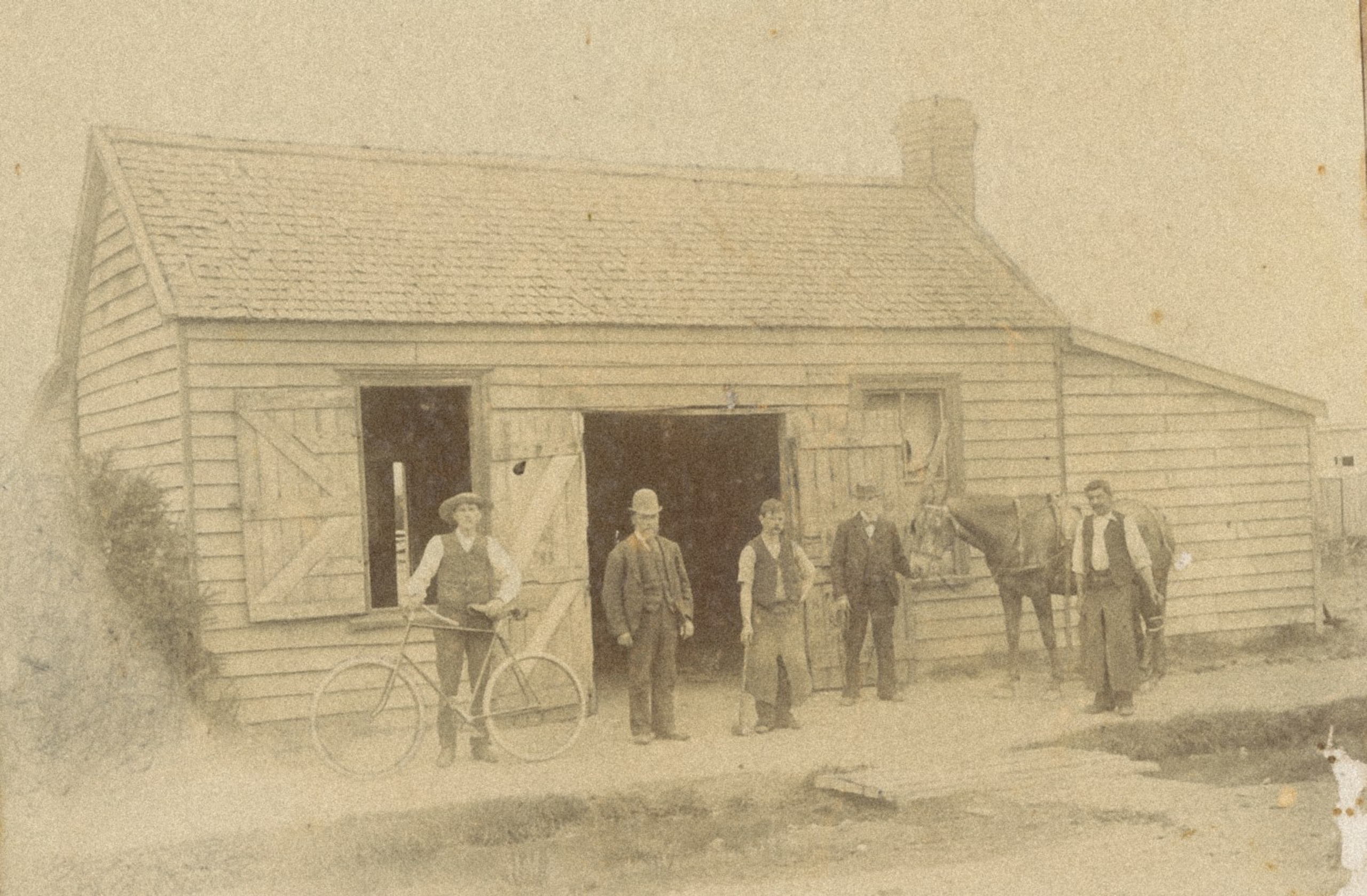 Early sepia photograph of a group of men standing outside John Staples’ weatherboard blacksmith shop in Tinwald, with a horse beside them.