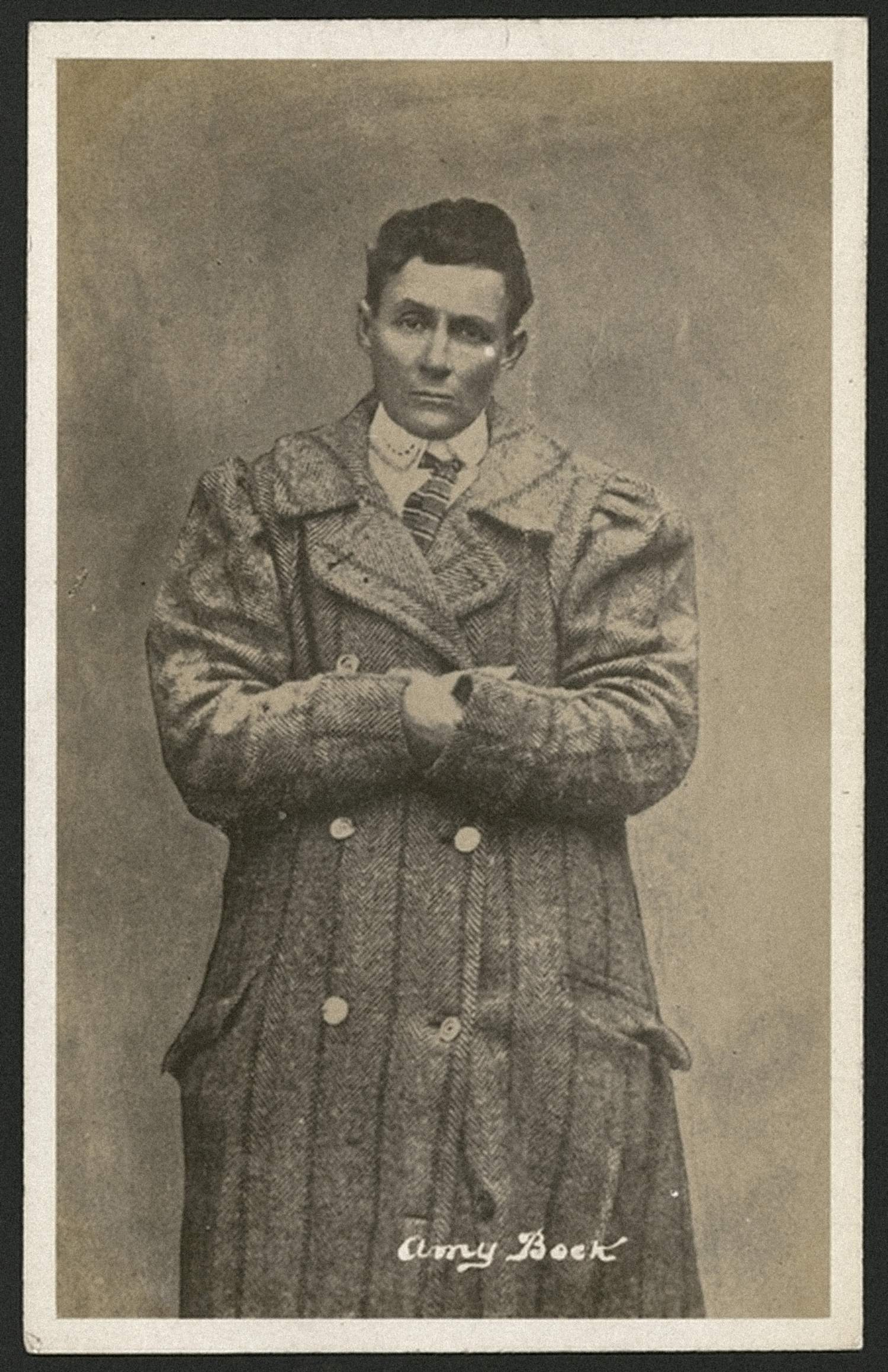 Postcard portrait of Amy Bock dressed in men’s clothing, standing in a long overcoat with a high collar.