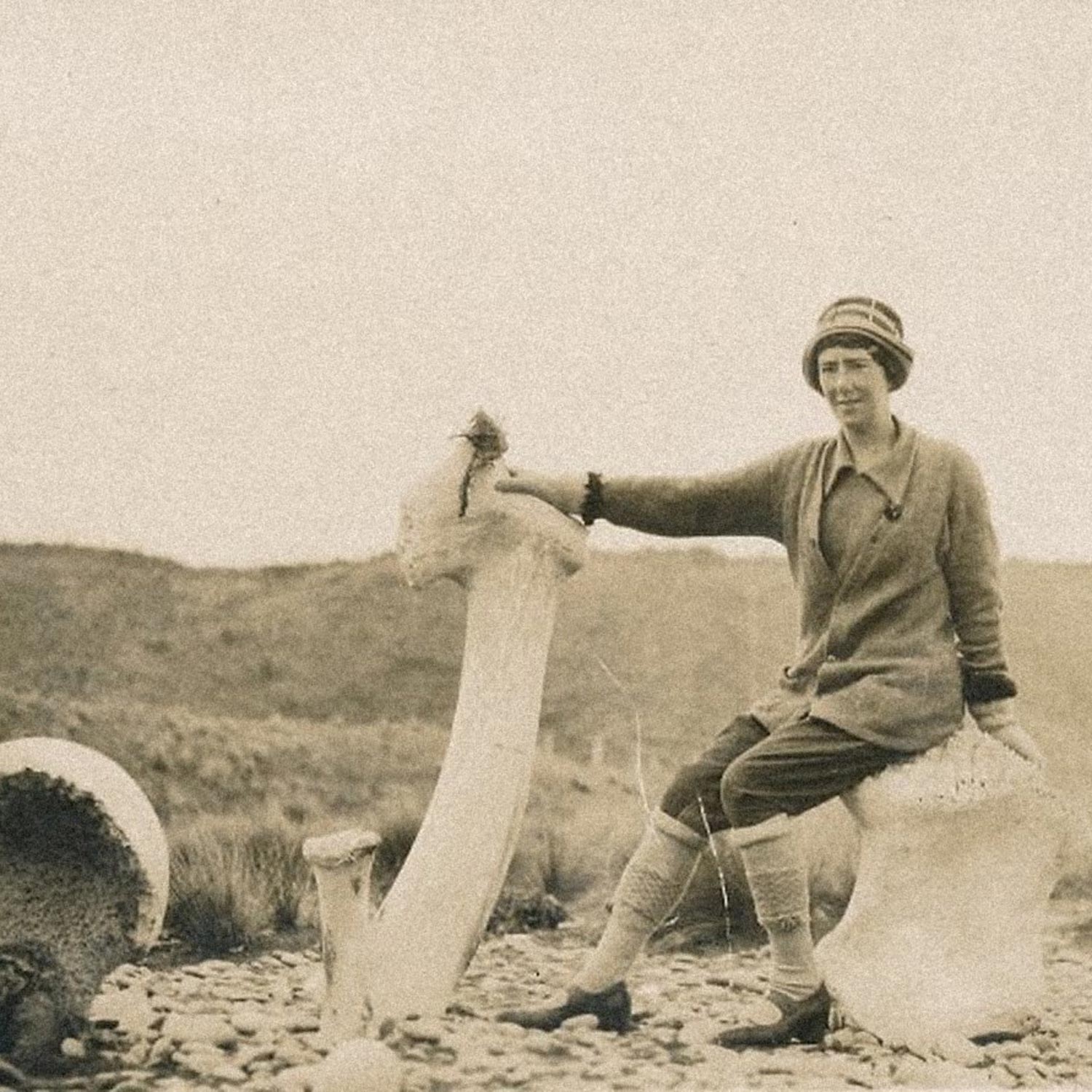 Sepia photograph of Daisy Mackie sitting on the shore at Whakanui beach beside large whale bones, including a tall jawbone standing upright.