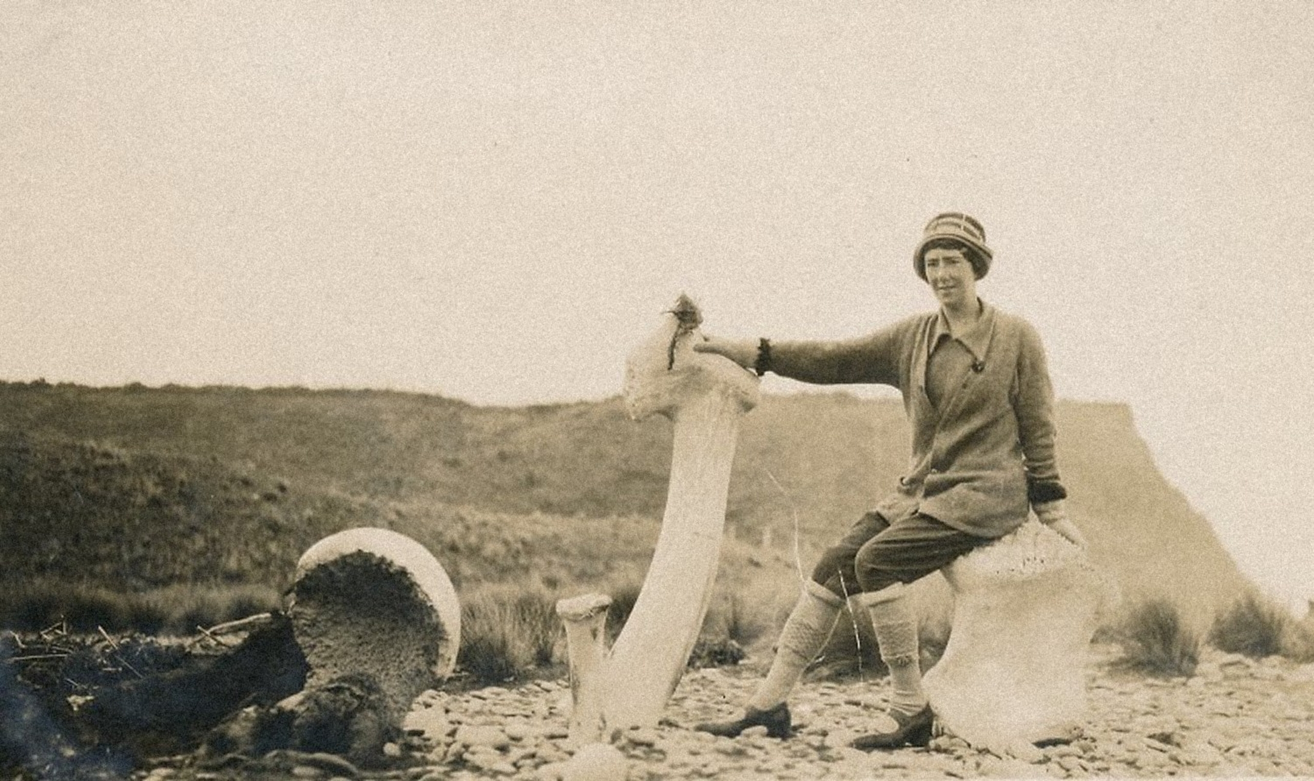Sepia photograph of Daisy Mackie sitting on the shore at Whakanui beach beside large whale bones, including a tall jawbone standing upright.