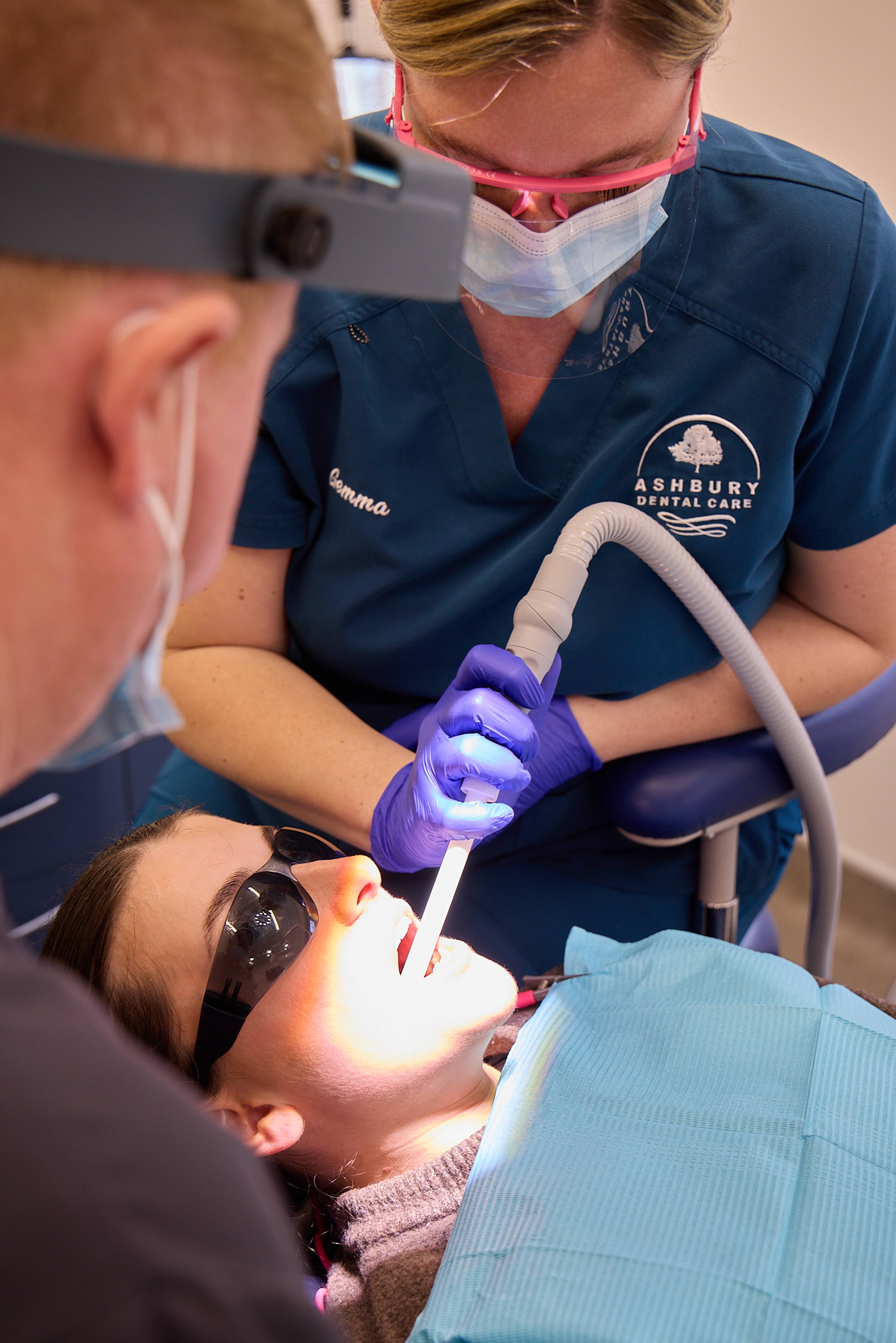 Nurse providing suction for patient wearing protective glasses in the dental chair with dentist out of focus in the frame