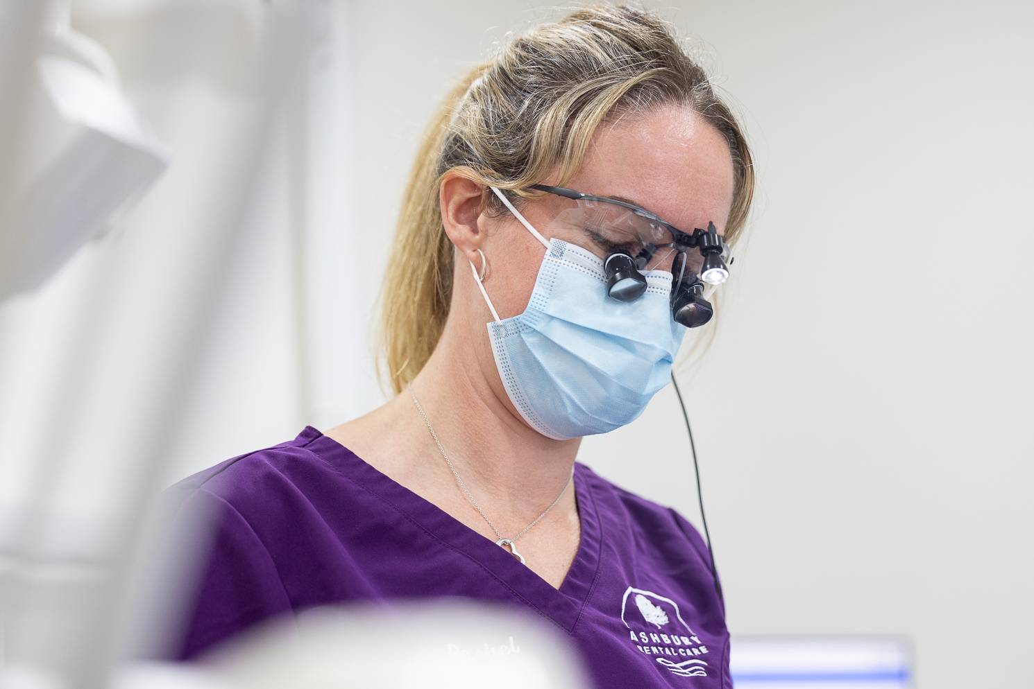 A dentist with a face mask and dental loupes looking down at a patient who is out of frame
