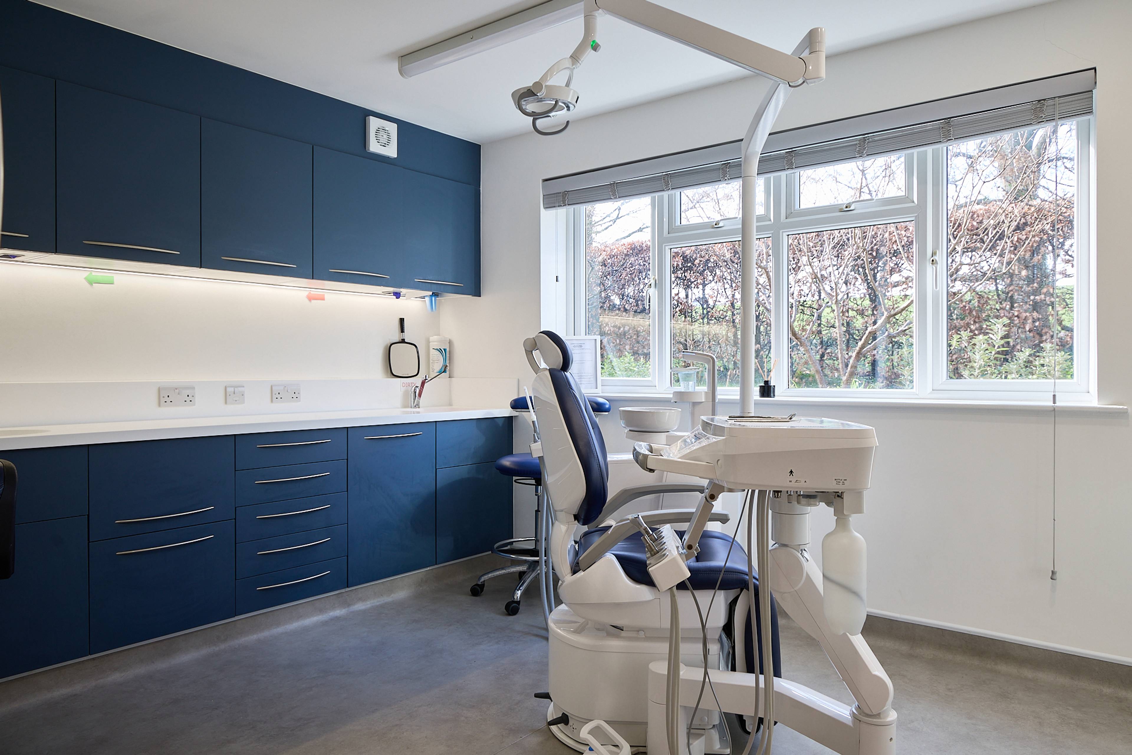 Modern clean dental surgery which a blue chair and cupboards and white walls with a big window