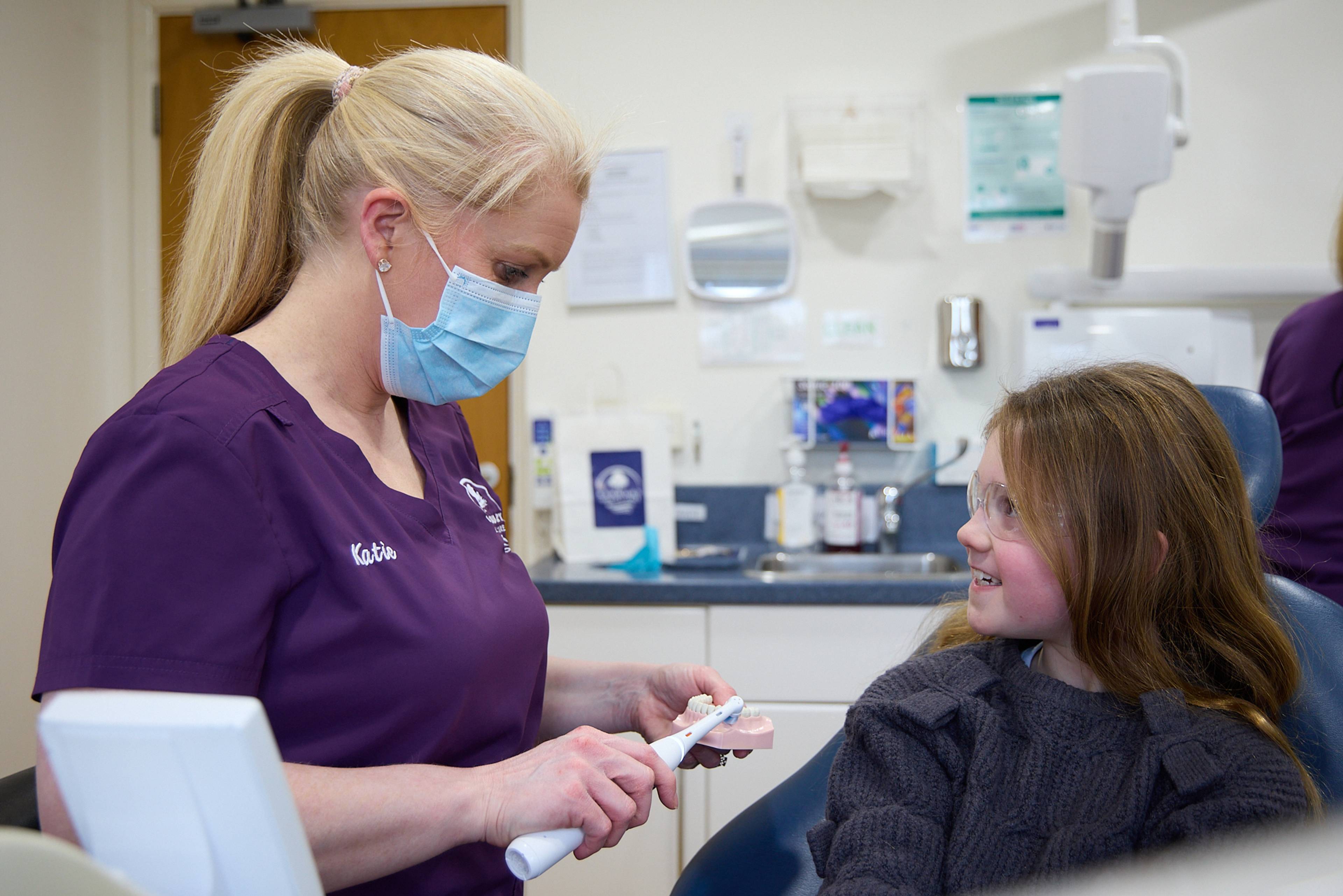 A masked hygienist at Ashbury Dental Care showing a child how to brush their teeth using a model