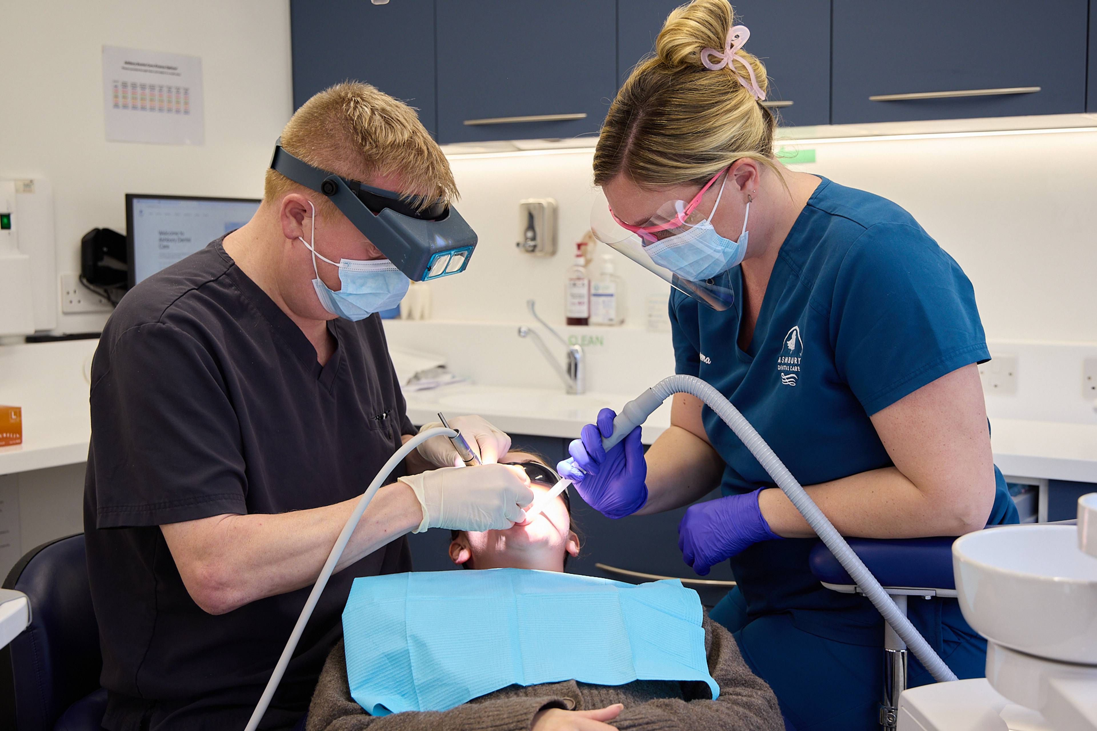 A masked dentist and nurse stand either side of the dental chair with suction and tools treating patient