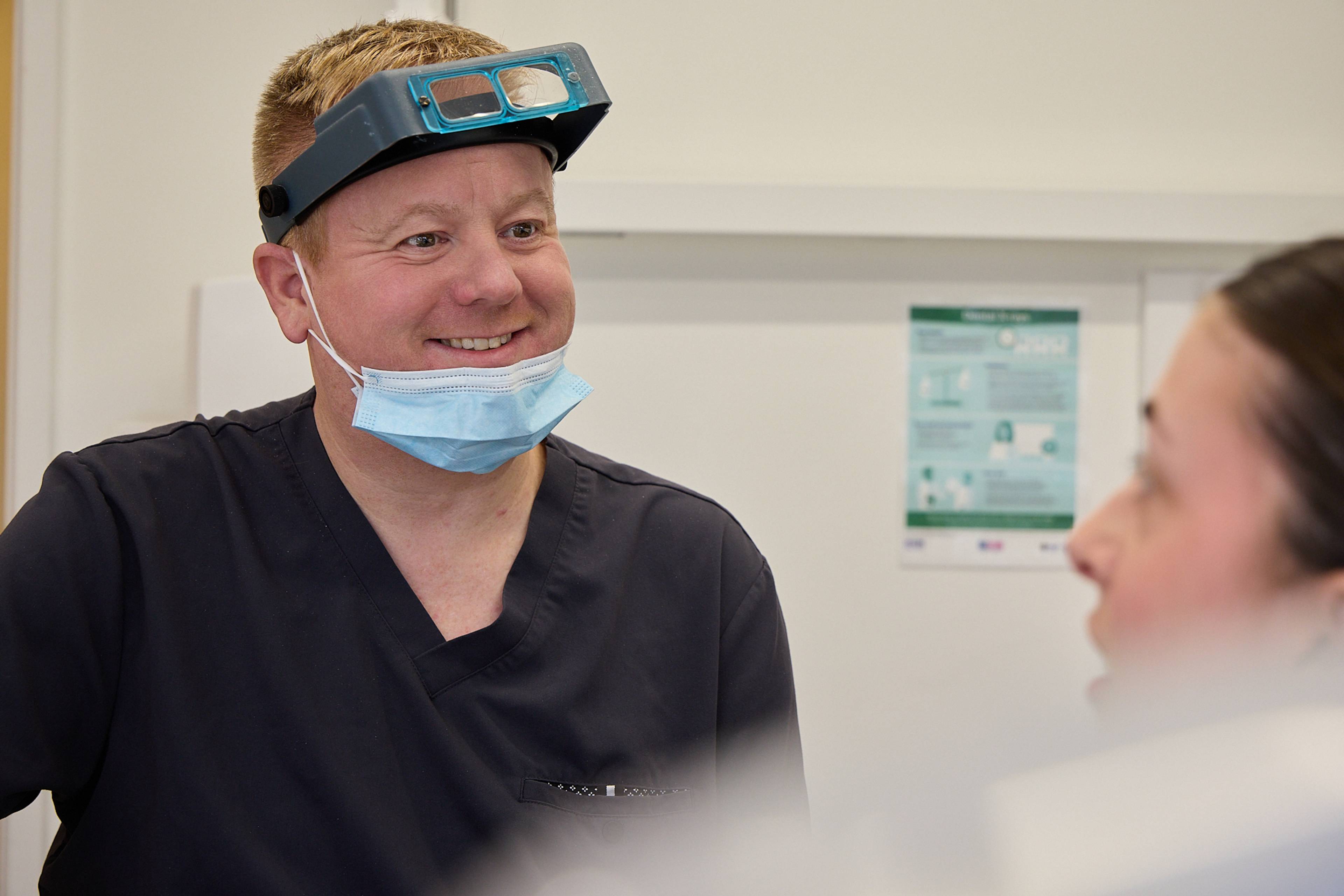 Smiling dentist wearing mask under their chin and loops on their head whilst speaking to patient in the dental chair