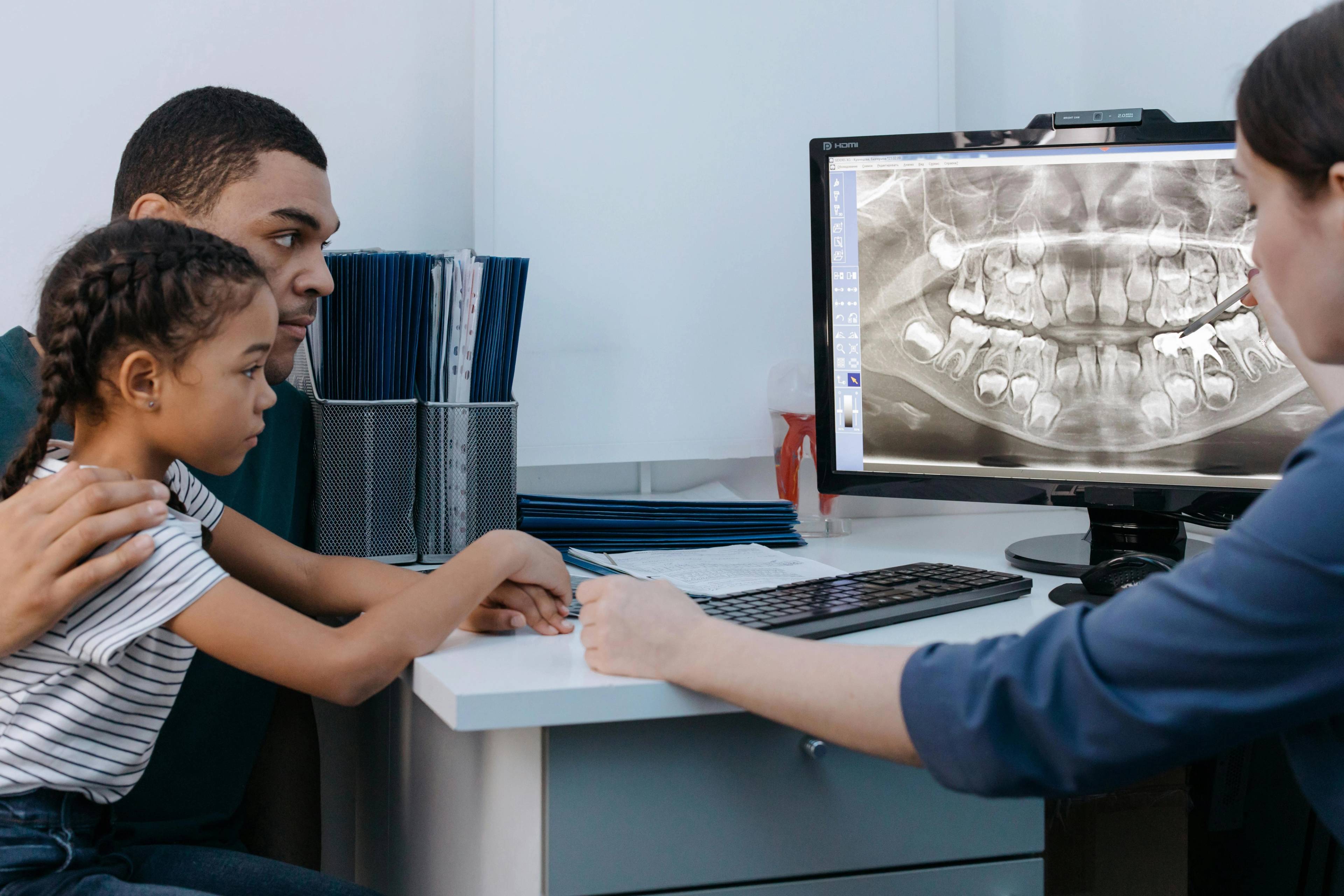 A dentist looks at an x-ray of a children's teeth on computer screen whilst patient and parent sit at the desk 