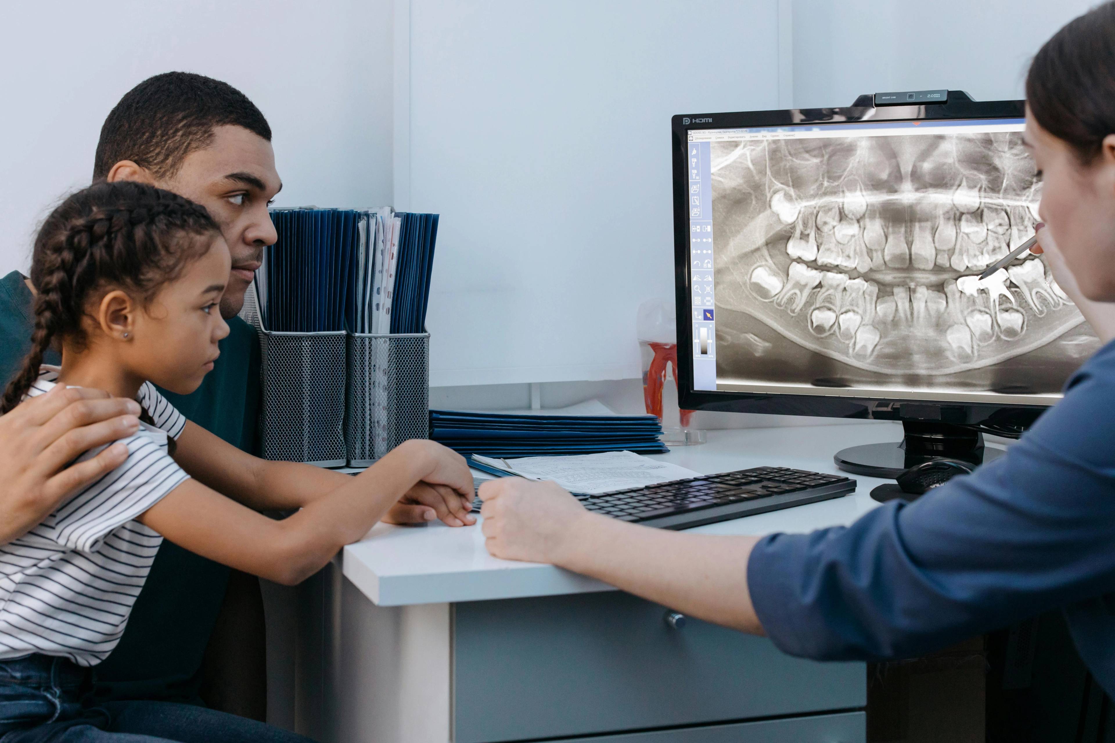 A dentist looks at an x-ray of a children's teeth on computer screen whilst patient and parent sit at the desk