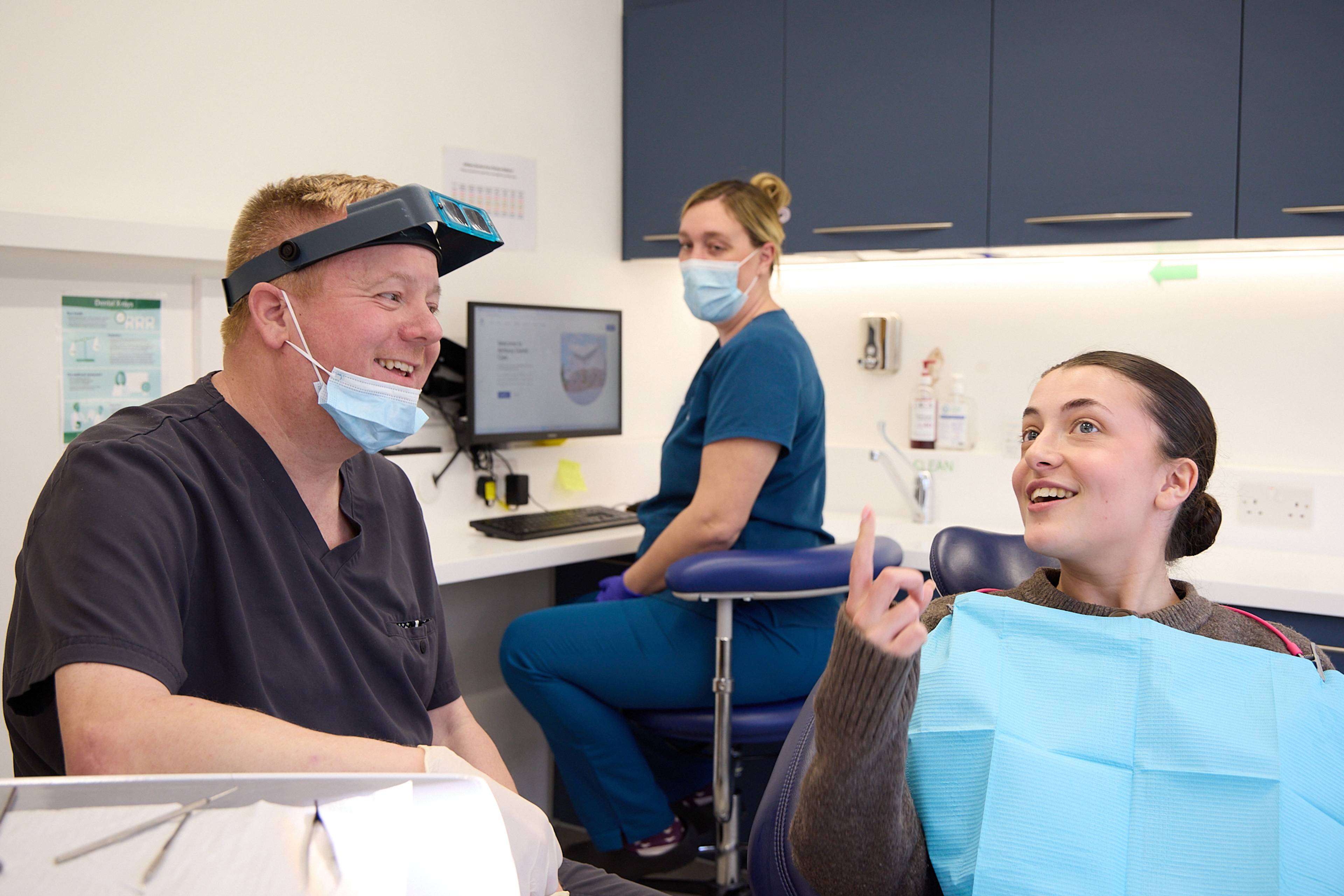 Dentist with mask and loops sat down talking to a patient in the chair at Ashbury Dental Care