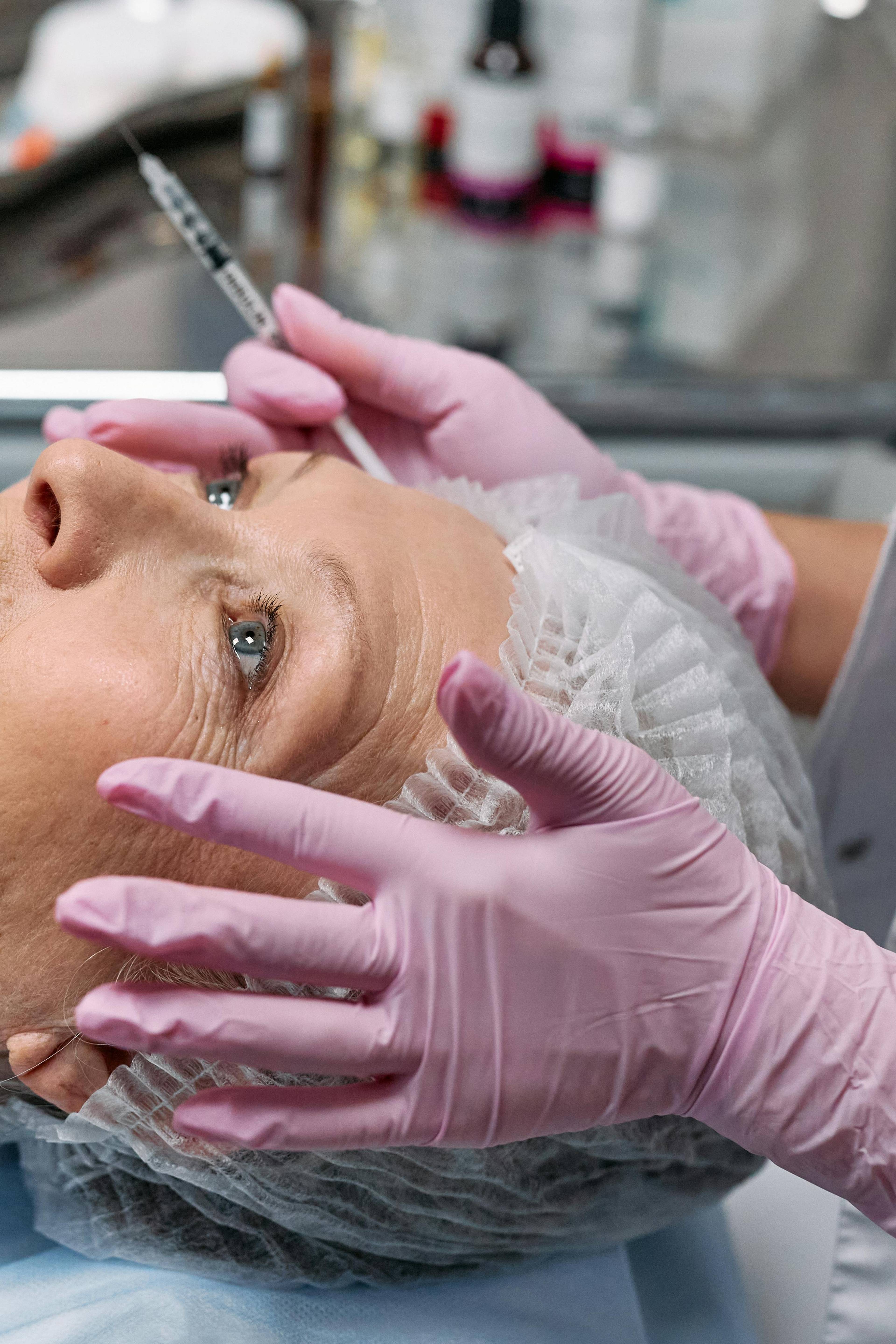 A patient about to receive a botox injection. An aesthetician's gloved hands hold the patient's face and a syringe.