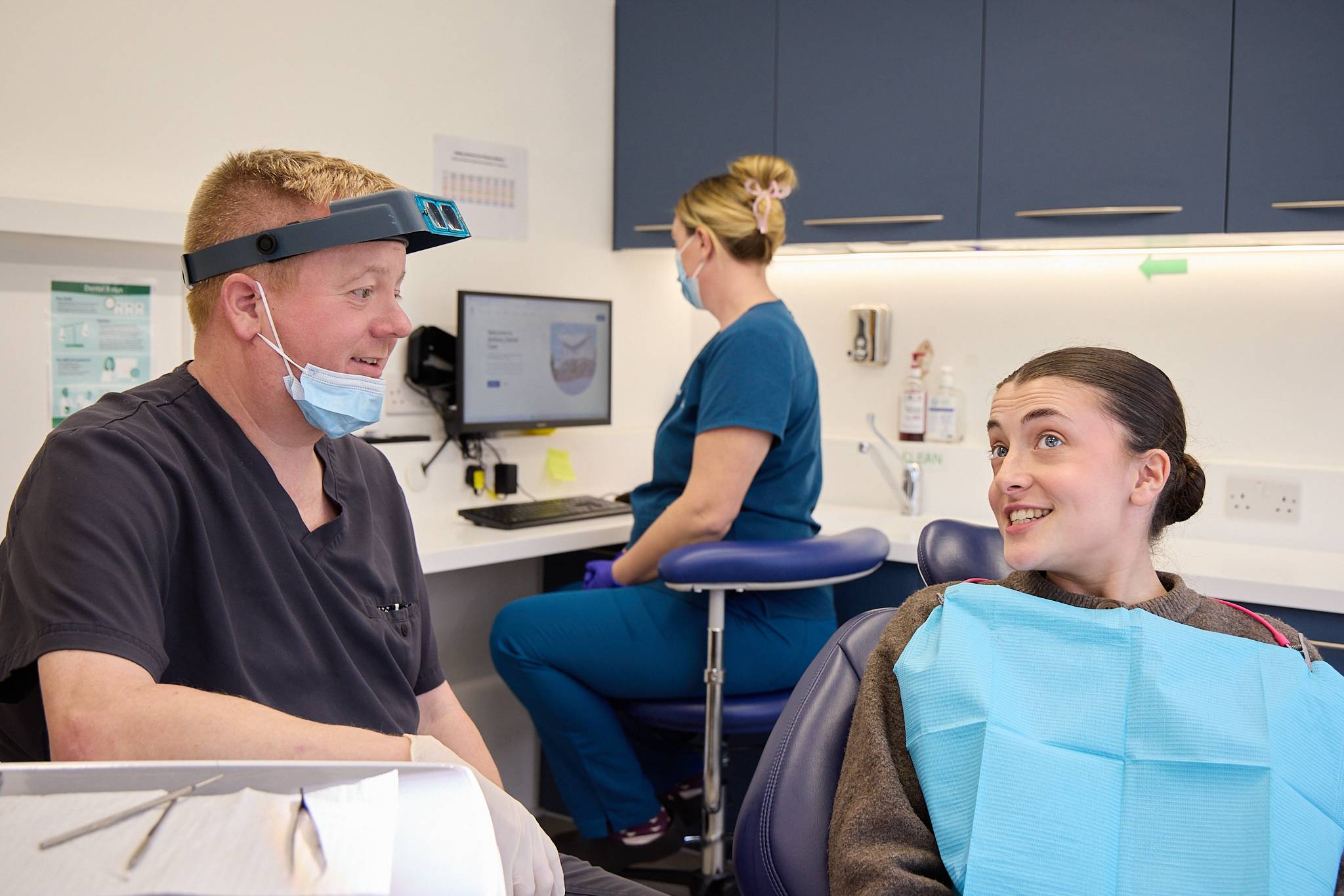 Dentists at Ashbury Dental Care wearing mask and loops, sat talking to patient in dental chair wearing blue bib