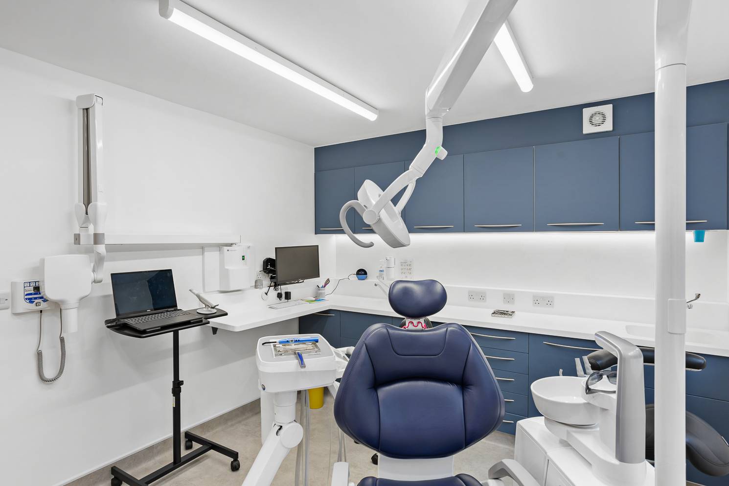 A picture of one of Ashbury Dental Care's treatment rooms, with a dark blue chair and cabinets and clean, well-lit, and welcoming surroundings.