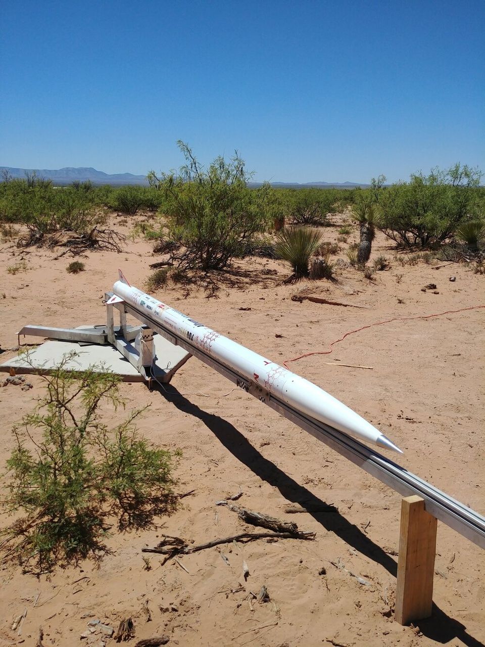 A white rocket with a pointed nose cone and small fins is resting horizontally on a launch rail in a dry, desert-like landscape. The rocket's body is covered with various small colored markings and logos. The launch rail, made of metal and wood, extends into the foreground, casting a shadow on the sandy ground. Sparse green bushes and other desert vegetation are scattered across the scene, with distant mountains visible under a clear blue sky.
