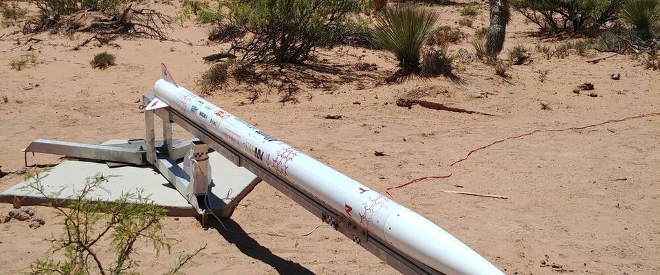 A white rocket with a pointed nose cone and small fins is resting horizontally on a launch rail in a dry, desert-like landscape. The rocket's body is covered with various small colored markings and logos. The launch rail, made of metal and wood, extends into the foreground, casting a shadow on the sandy ground. Sparse green bushes and other desert vegetation are scattered across the scene, with distant mountains visible under a clear blue sky.