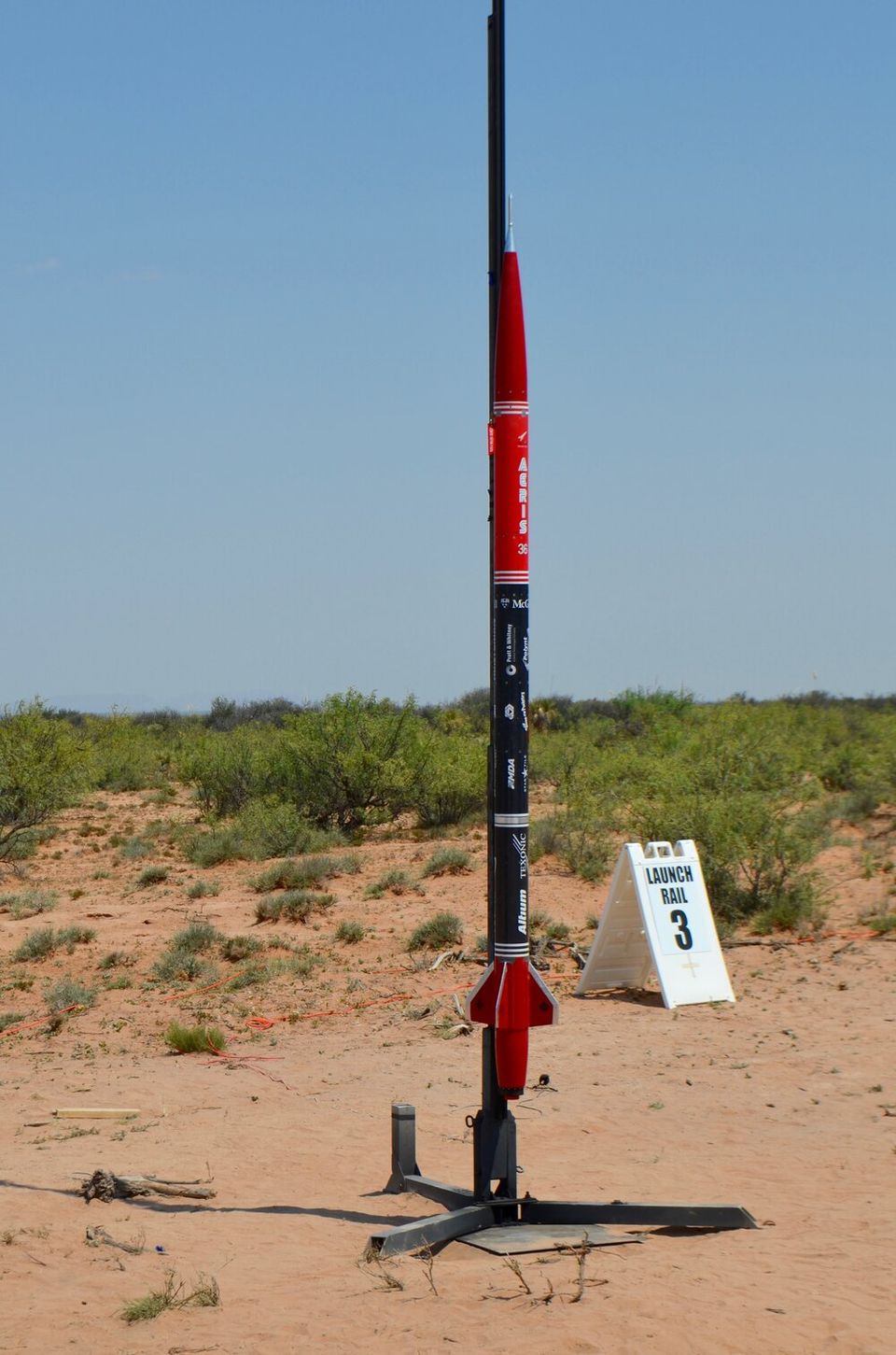 A tall, slender rocket, primarily black with a red nose cone and red fins, stands upright on a launch rail in a desert-like landscape. The rocket has "ARIES" and other text visible on its side. To the right of the rocket, a white A-frame sign reads "LAUNCH RAIL 3". The ground is sandy with sparse, low-lying vegetation, and the sky above is clear blue.