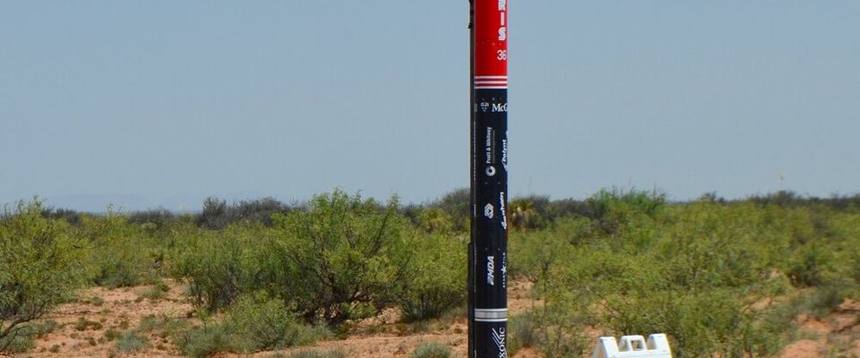 A tall, slender rocket, primarily black with a red nose cone and red fins, stands upright on a launch rail in a desert-like landscape. The rocket has "ARIES" and other text visible on its side. To the right of the rocket, a white A-frame sign reads "LAUNCH RAIL 3". The ground is sandy with sparse, low-lying vegetation, and the sky above is clear blue.