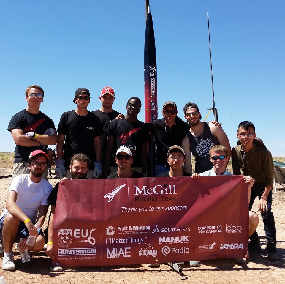 A group of approximately 15 young adults, identified as the McGill Rocket Team, are gathered outdoors on what appears to be a dry, unpaved field under a clear blue sky. They are posing for a photo with a large rocket, predominantly black with some red accents, standing vertically behind them. The rocket has "McGILL" and "SAINT-JEAN-SUR-RICHELIEU" visible on its side. In front of the group, held by several team members, is a large red banner displaying "McGill Rocket Team" and "Thank you to our sponsors" along with various sponsor logos. The team members are dressed in casual attire, including t-shirts and shorts, and some are wearing hats and sunglasses. The overall impression is one of accomplishment and teamwork in an outdoor, possibly remote, setting.