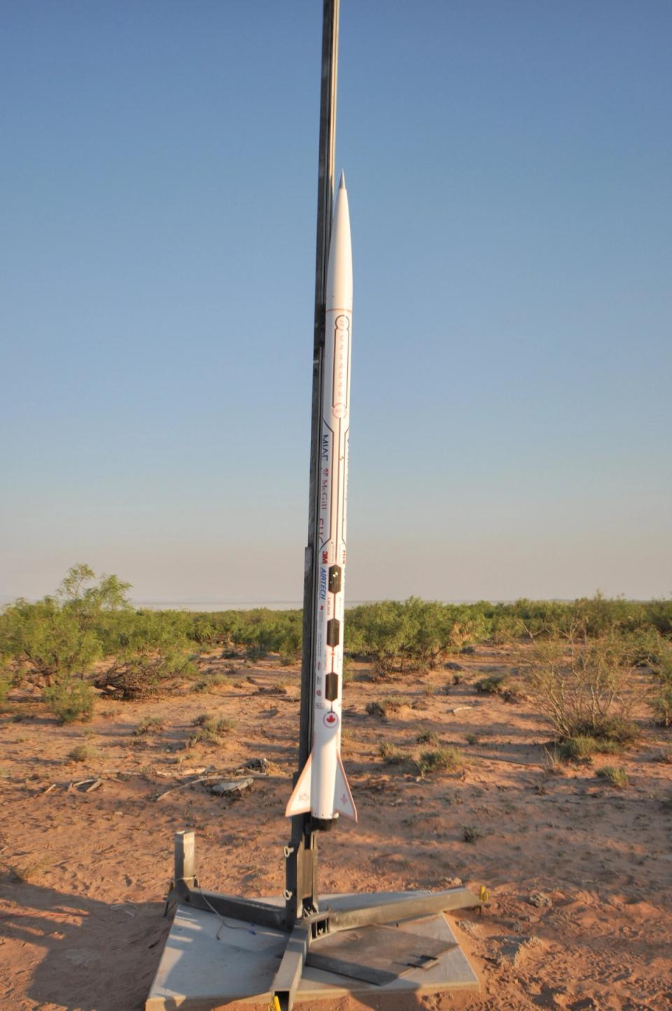 A tall, white rocket, Project Caladan, stands upright on a launchpad in a dry, open field under a clear blue sky. The rocket has a Canadian maple leaf logo near its base and various other markings and sponsors along its body. The ground around the launchpad is reddish-brown dirt with sparse green and brown bushes.
