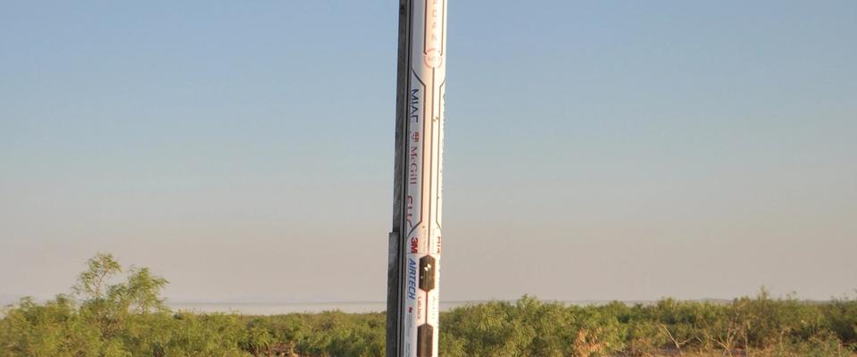 A tall, white rocket, Project Caladan, stands upright on a launchpad in a dry, open field under a clear blue sky. The rocket has a Canadian maple leaf logo near its base and various other markings and sponsors along its body. The ground around the launchpad is reddish-brown dirt with sparse green and brown bushes.
