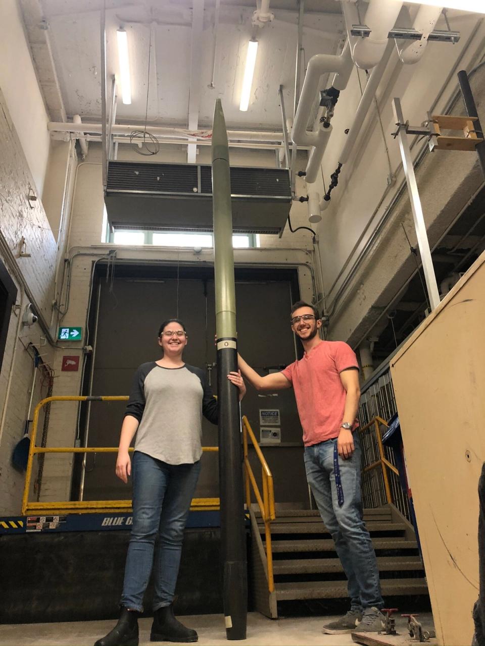 Two smiling McGill Rocket Team members, a woman and a man, stand proudly beside their tall, two-toned Project Aramis rocket in a lab setting.