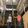 Two smiling McGill Rocket Team members, a woman and a man, stand proudly beside their tall, two-toned Project Aramis rocket in a lab setting.