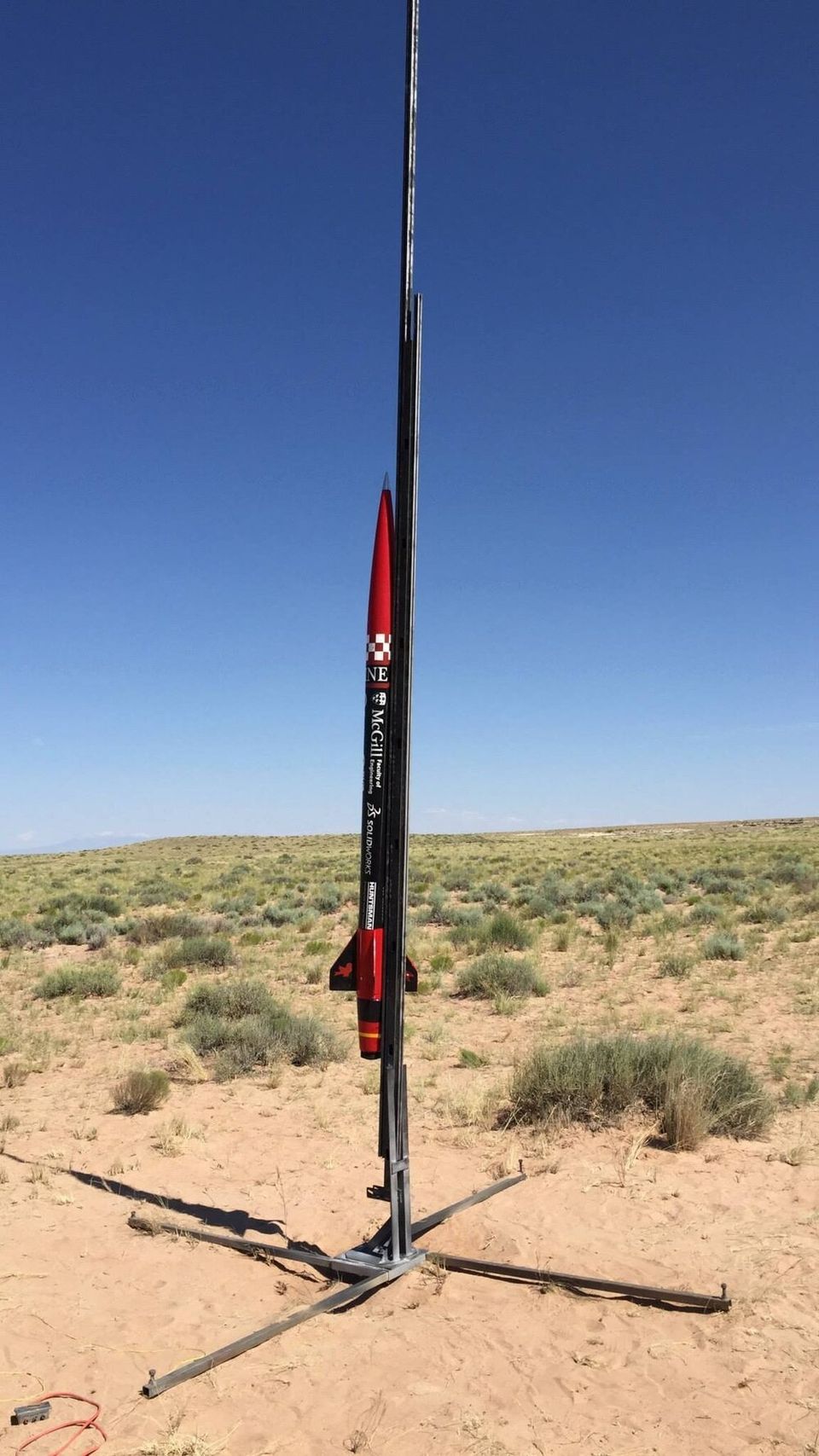 A slender, red and black rocket, emblazoned with "McGill" and other text, stands vertically on a launch rail in a dry, open field under a clear blue sky.