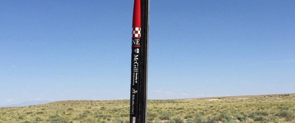 A slender, red and black rocket, emblazoned with "McGill" and other text, stands vertically on a launch rail in a dry, open field under a clear blue sky.