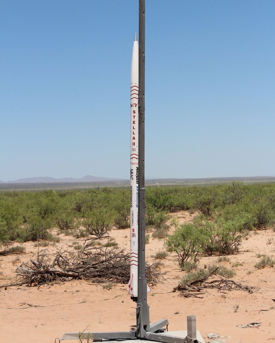 A tall, slender white rocket with red markings, including the name "STELLA" written vertically, stands upright on a launch rail in a desert landscape. The ground is sandy with sparse green bushes and some dried brush. In the background, a flat horizon separates the desert from a clear, bright blue sky.