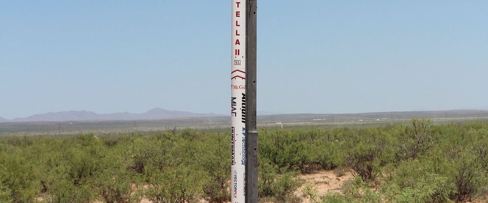 A tall, slender white rocket with red markings, including the name "STELLA" written vertically, stands upright on a launch rail in a desert landscape. The ground is sandy with sparse green bushes and some dried brush. In the background, a flat horizon separates the desert from a clear, bright blue sky.