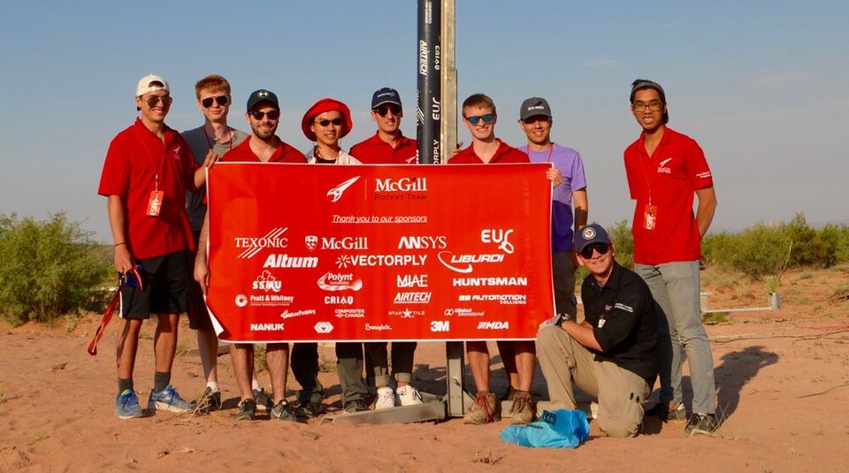 A group of nine team members, are posing for a photo in an arid, outdoor environment. Behind them stands a tall, slender rocket, predominantly black with a white nose cone, on a launch rail. The team members are holding a large red banner that reads "McGill Rocket Team" and "Thank you to our sponsors," along with various company logos. The individuals are casually dressed, some wearing hats and sunglasses, and they appear to be in good spirits, celebrating their work with the rocket. The sky above is clear and blue.