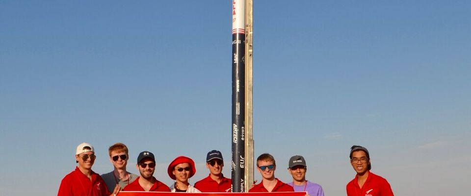 A group of nine team members, are posing for a photo in an arid, outdoor environment. Behind them stands a tall, slender rocket, predominantly black with a white nose cone, on a launch rail. The team members are holding a large red banner that reads "McGill Rocket Team" and "Thank you to our sponsors," along with various company logos. The individuals are casually dressed, some wearing hats and sunglasses, and they appear to be in good spirits, celebrating their work with the rocket. The sky above is clear and blue.