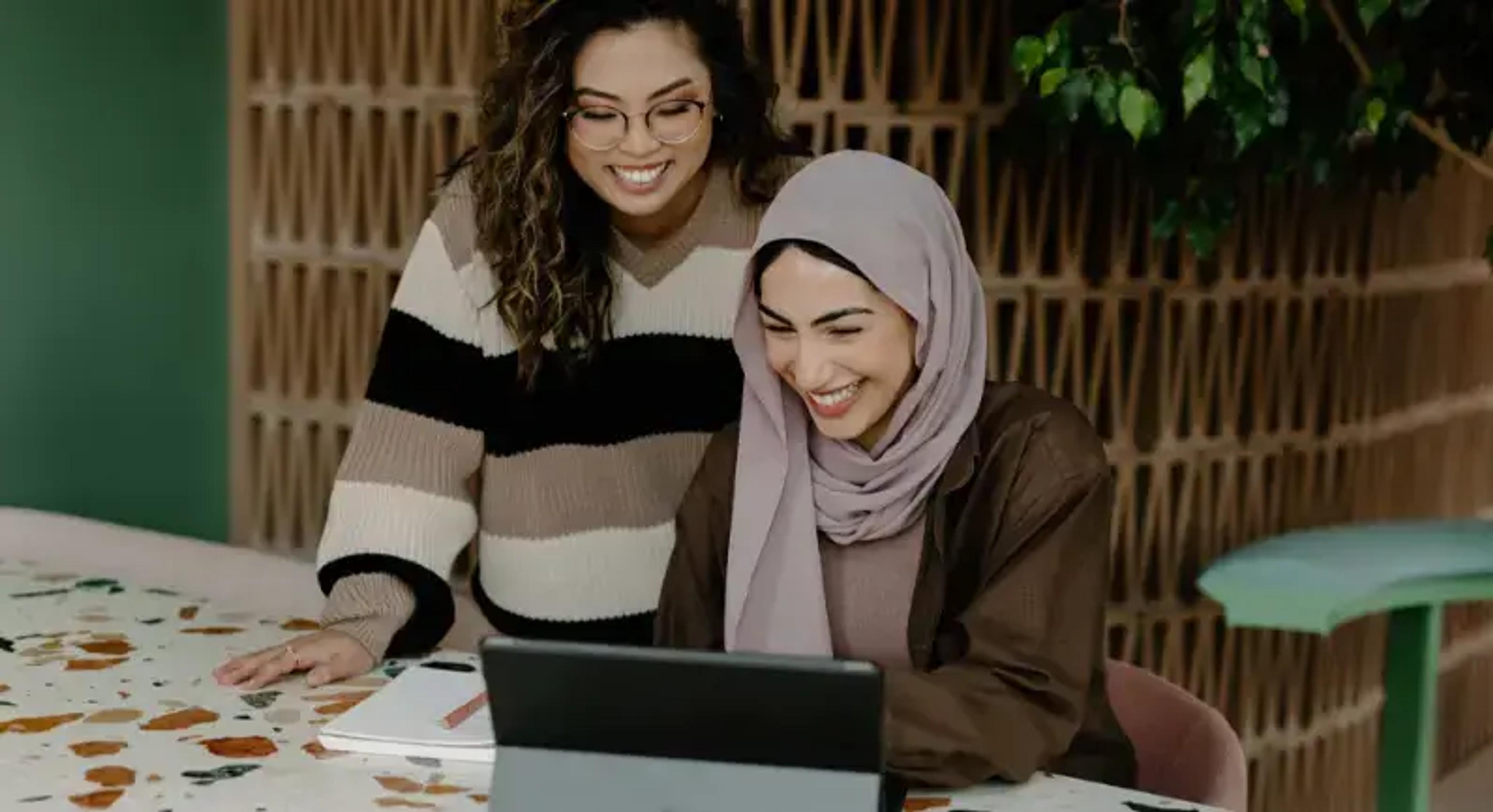 Twee lachende vrouwen die samen aan een laptop werken aan een kleurrijke tafel