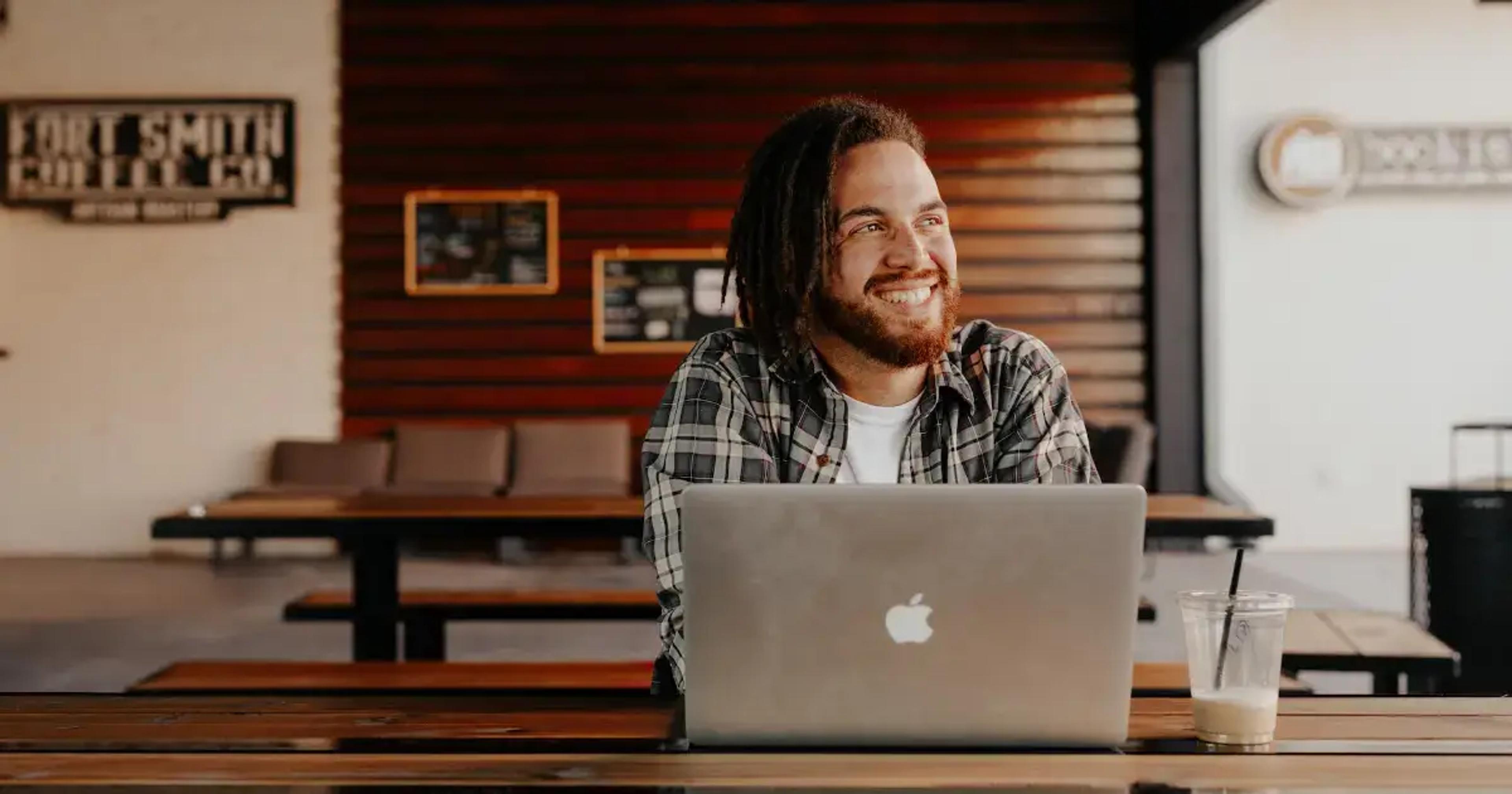 Smilende person arbejder på MacBook ved træbord i moderne café