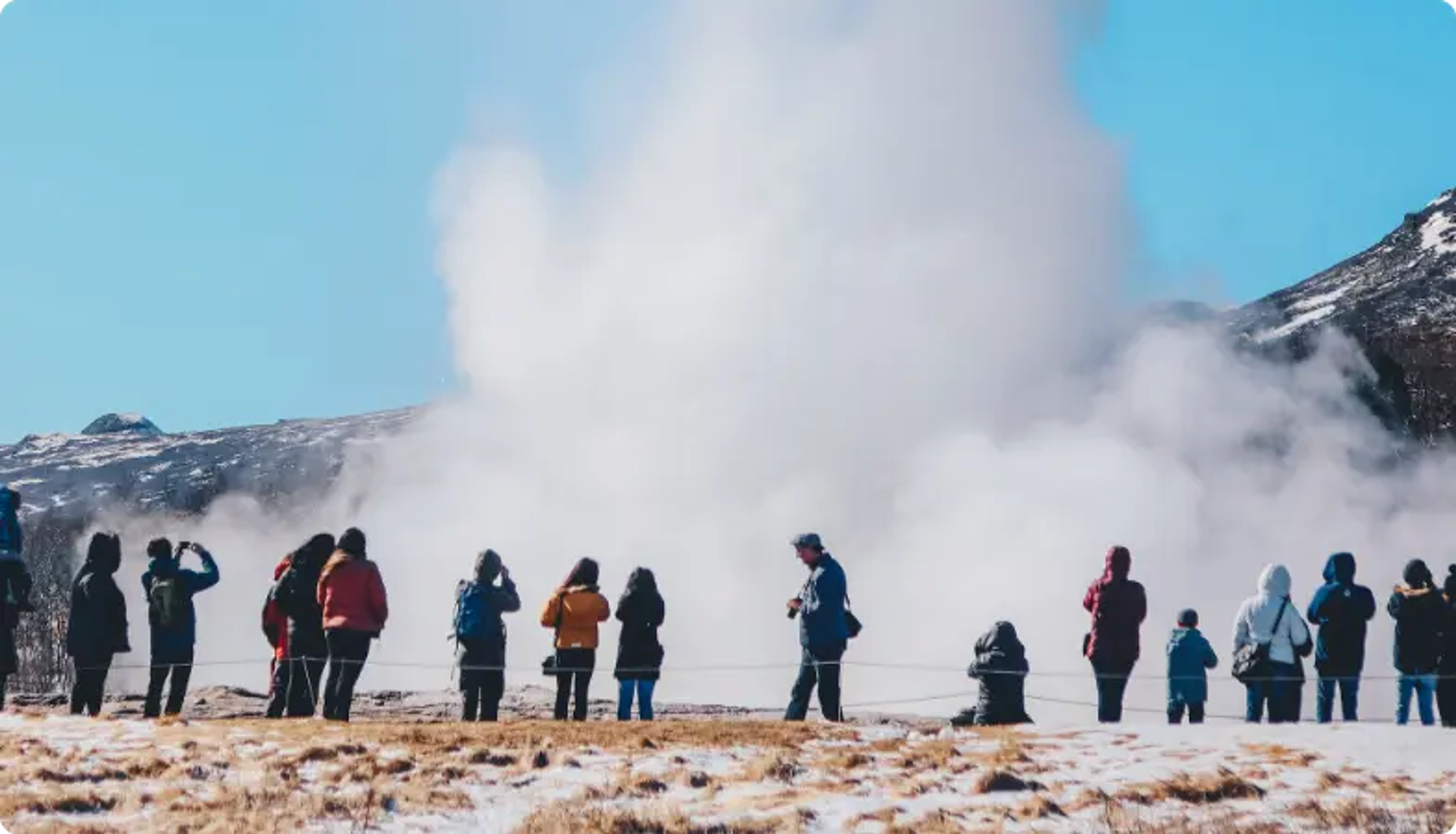 Touristen beobachten einen großen Geysir-Ausbruch vor einer schneebedeckten Berglandschaft