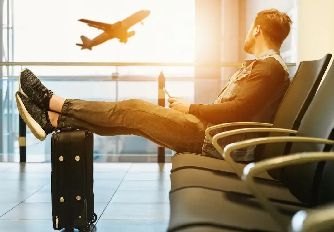 Utazó várakozik a repülőtéren, háttérben felszálló repülőgép, közelben poggyász