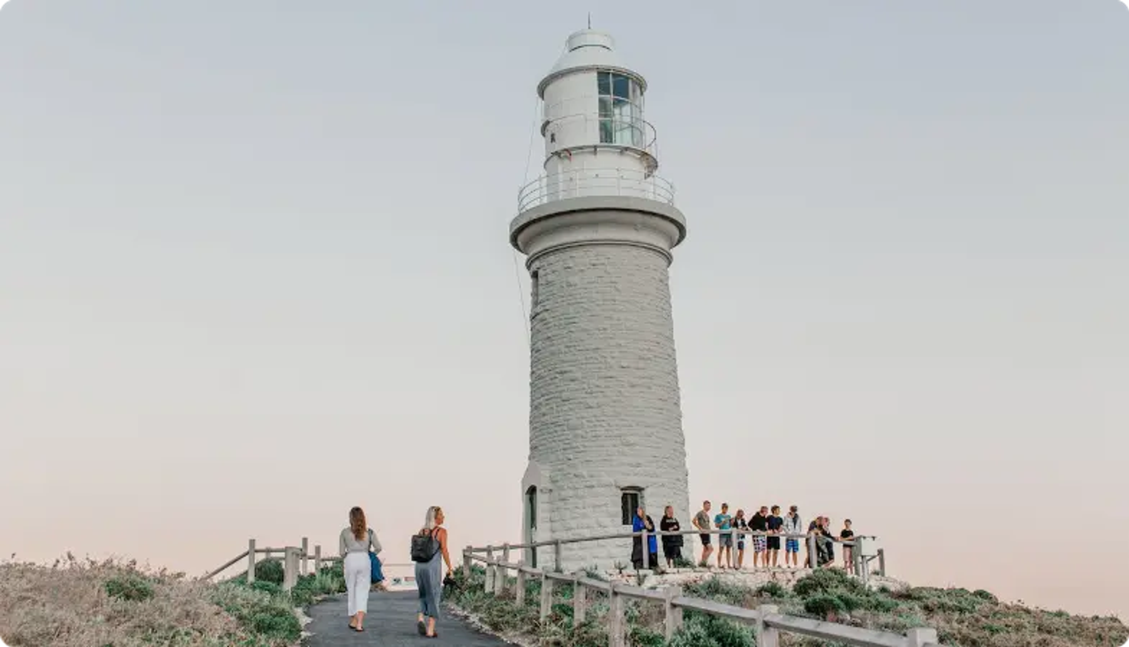 White lighthouse with tourists on wooden walkway overlooking coastal landscape