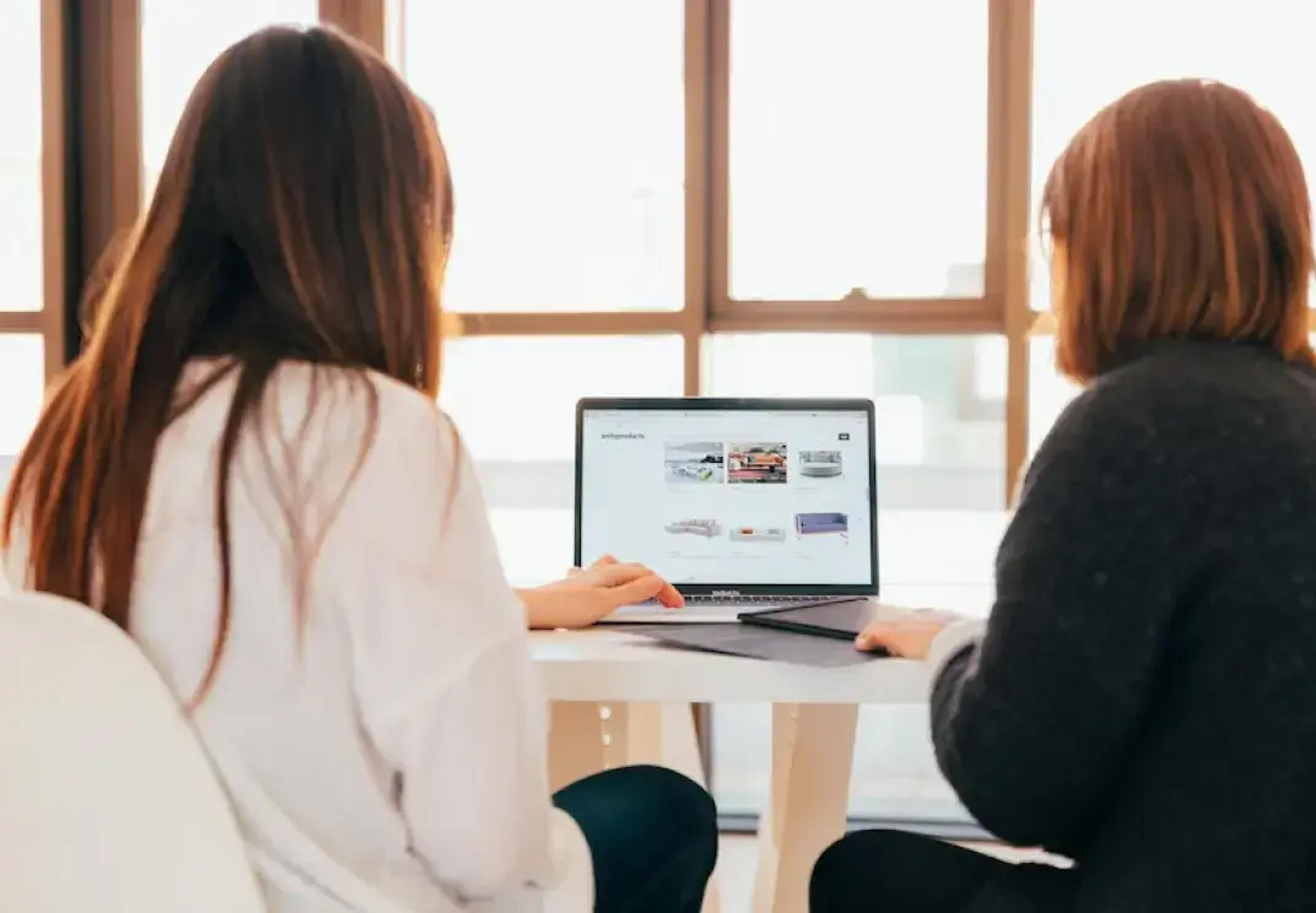 Két ember laptopot néz, amelyen egy weboldal van megnyitva, világos ablakok közelében