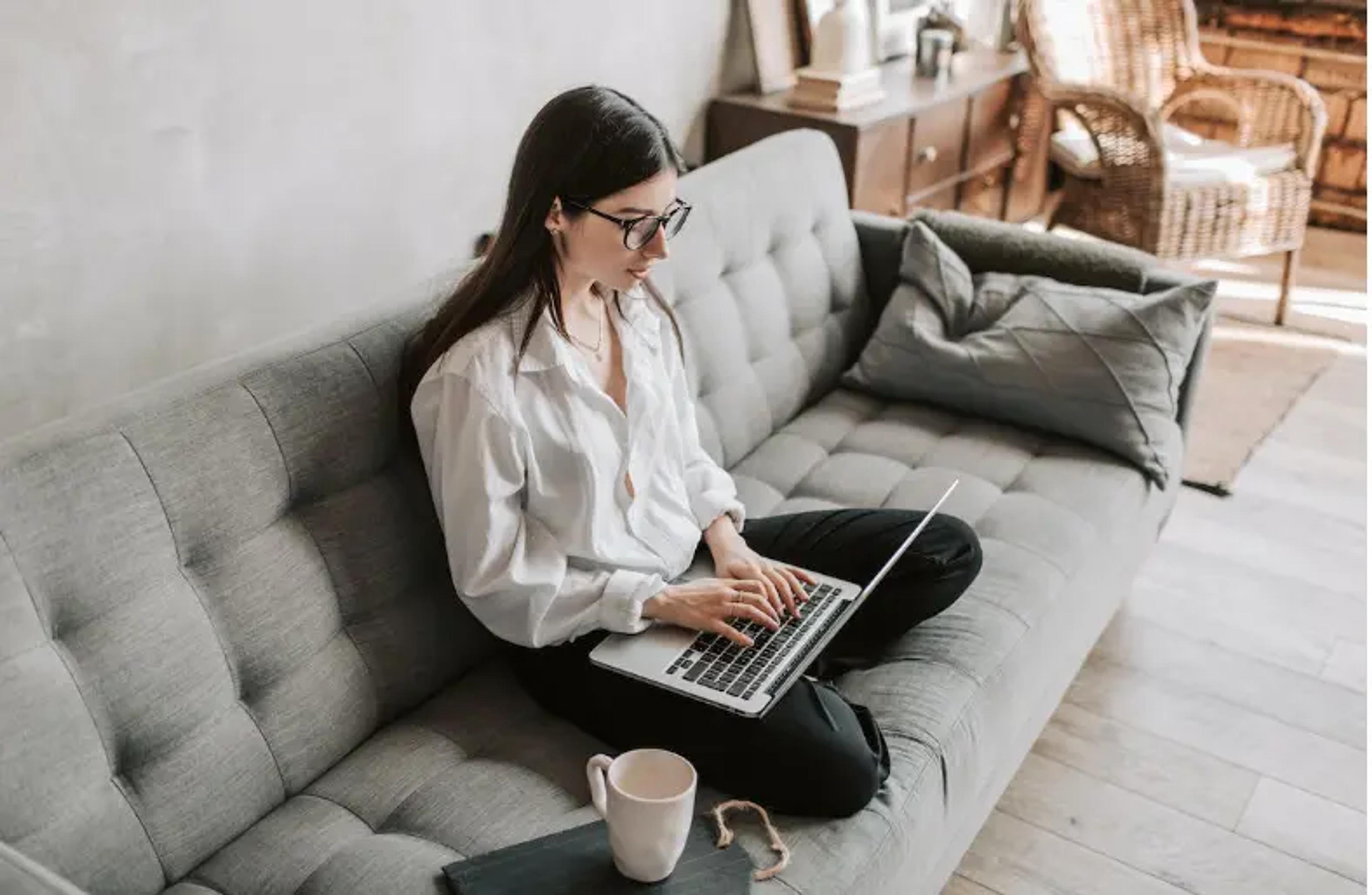 Mulher trabalhando no laptop sentada em um sofá aconchegante com uma caneca de café ao lado