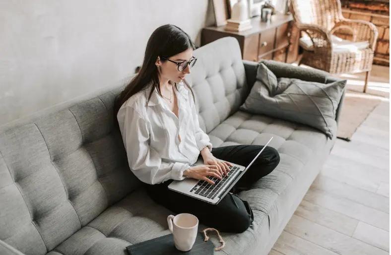 Vrouw die op laptop werkt zittend op een gezellige bank met koffiemok in de buurt