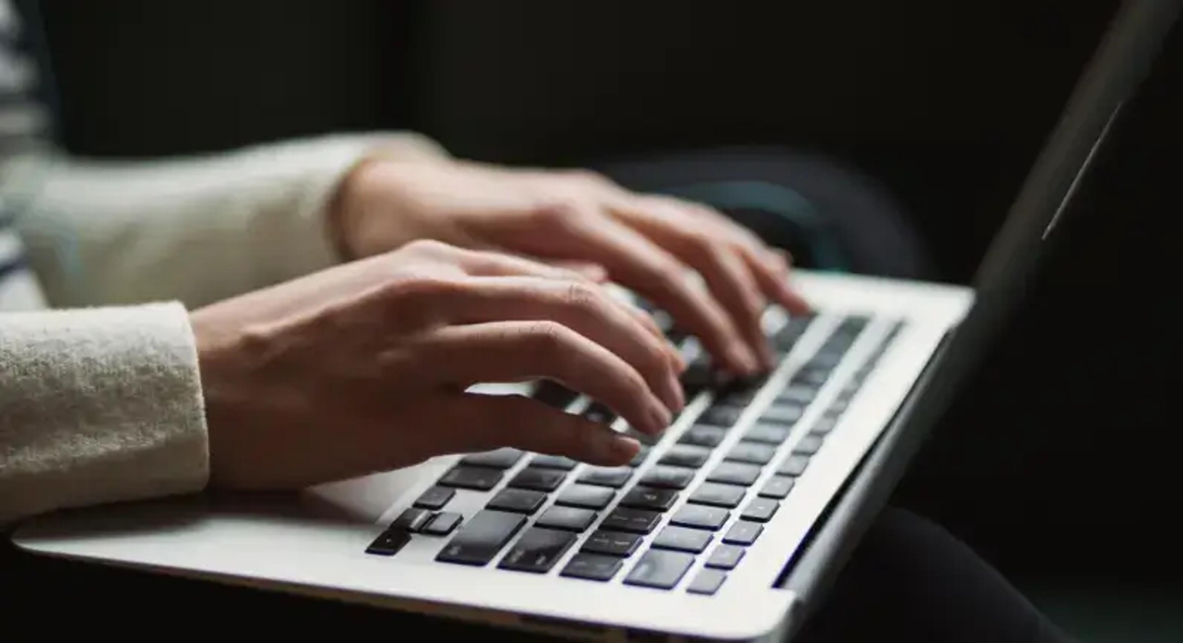 Kezek gépelnek egy laptop billentyűzetén egy félhomályos környezetben