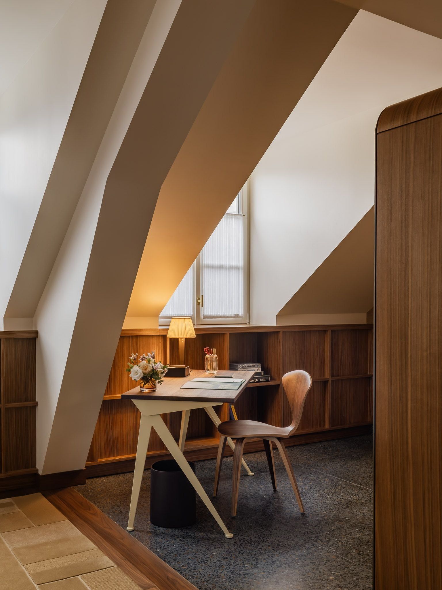 Desk and curved wooden chair tucked beneath sloping white beams, with a lit table lamp, flowers and a narrow window above wood paneling.