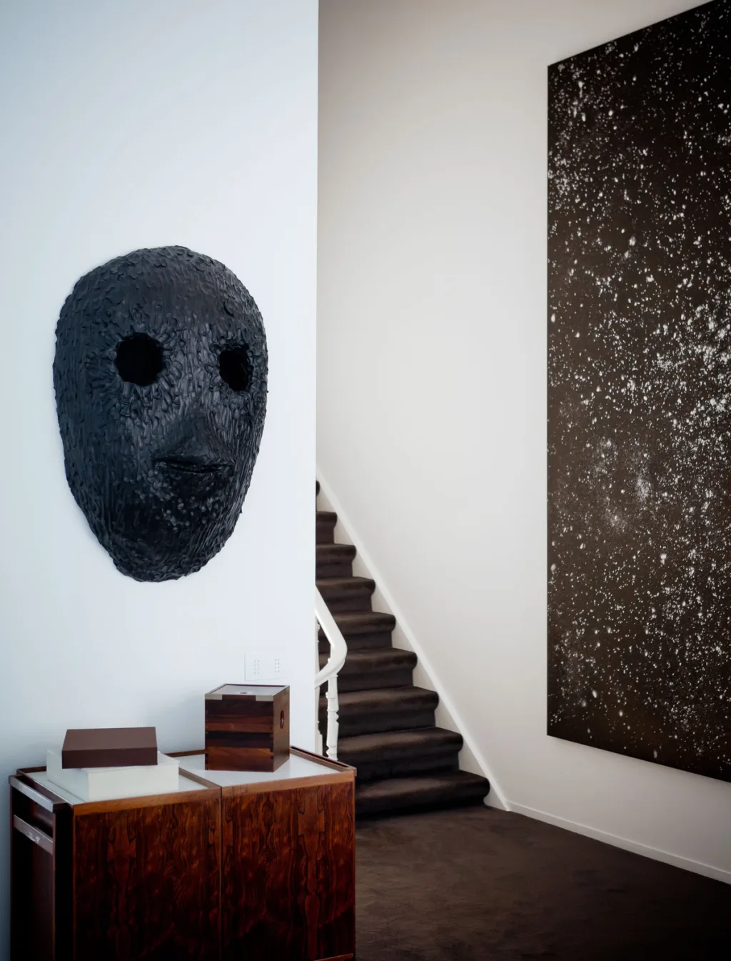 Dark sculptural mask and tall speckled artwork beside a staircase in the James Van Damme series