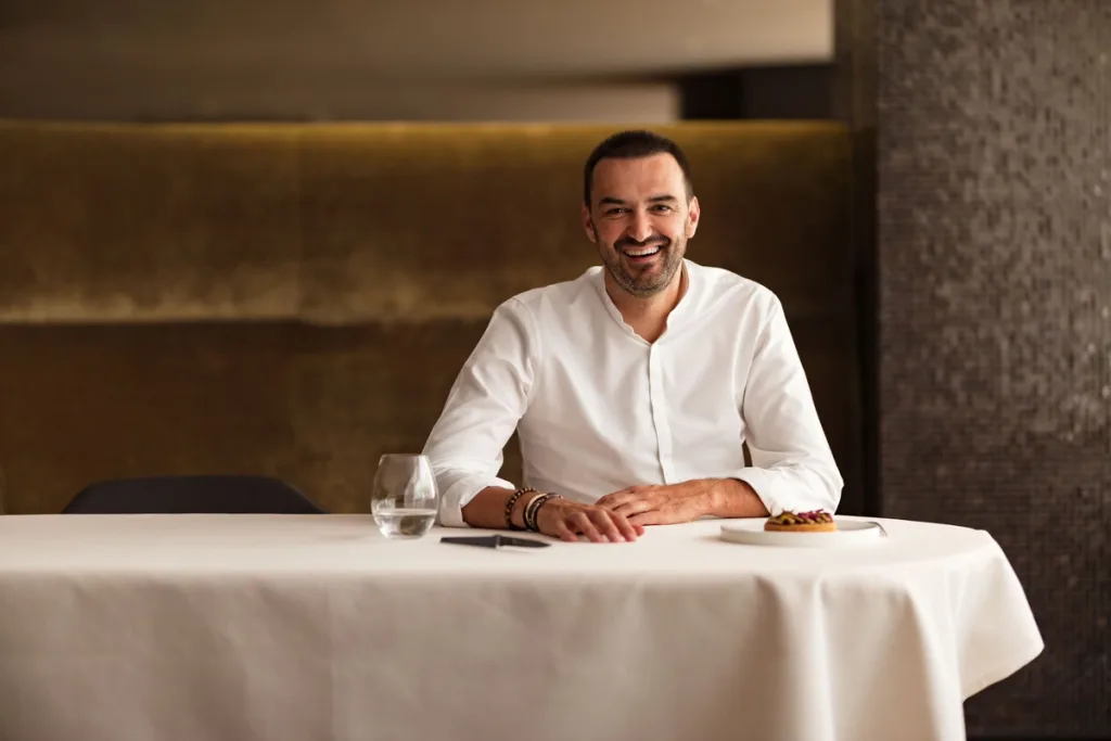 Smiling chef seated behind a white tablecloth and a small dessert for Cyril Lignac’s Saisons cookbook