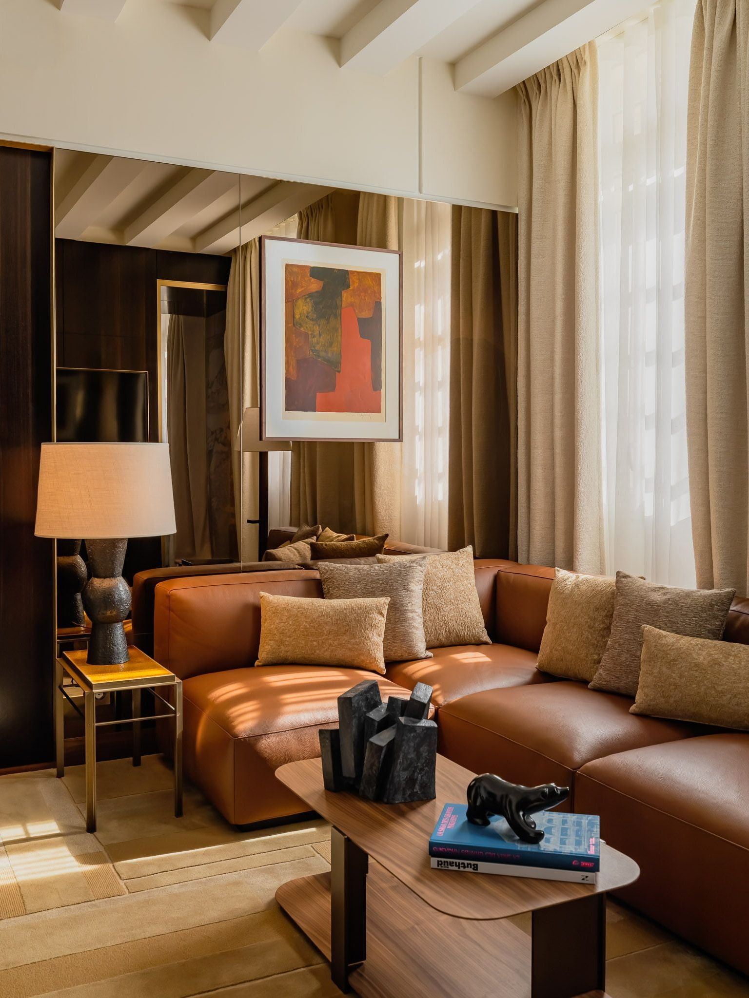 Caramel leather corner sofa beside tall curtained windows, with a mirrored wall, framed abstract art and nested coffee tables holding books and sculptures.