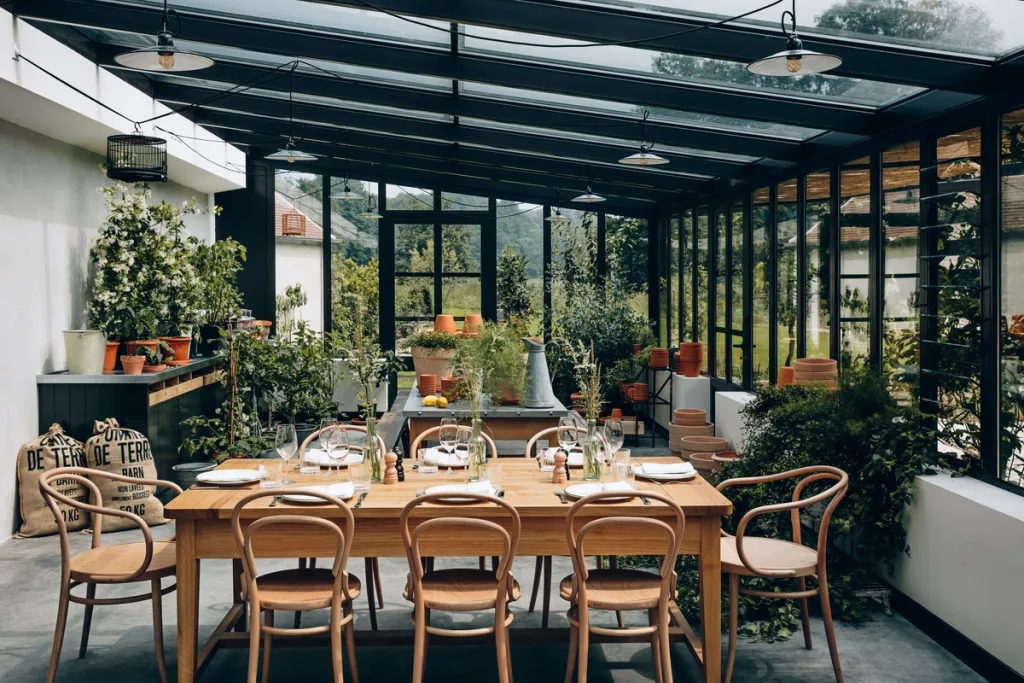 Long wooden table set for dinner in a glass-roofed conservatory, Le Barn hotel